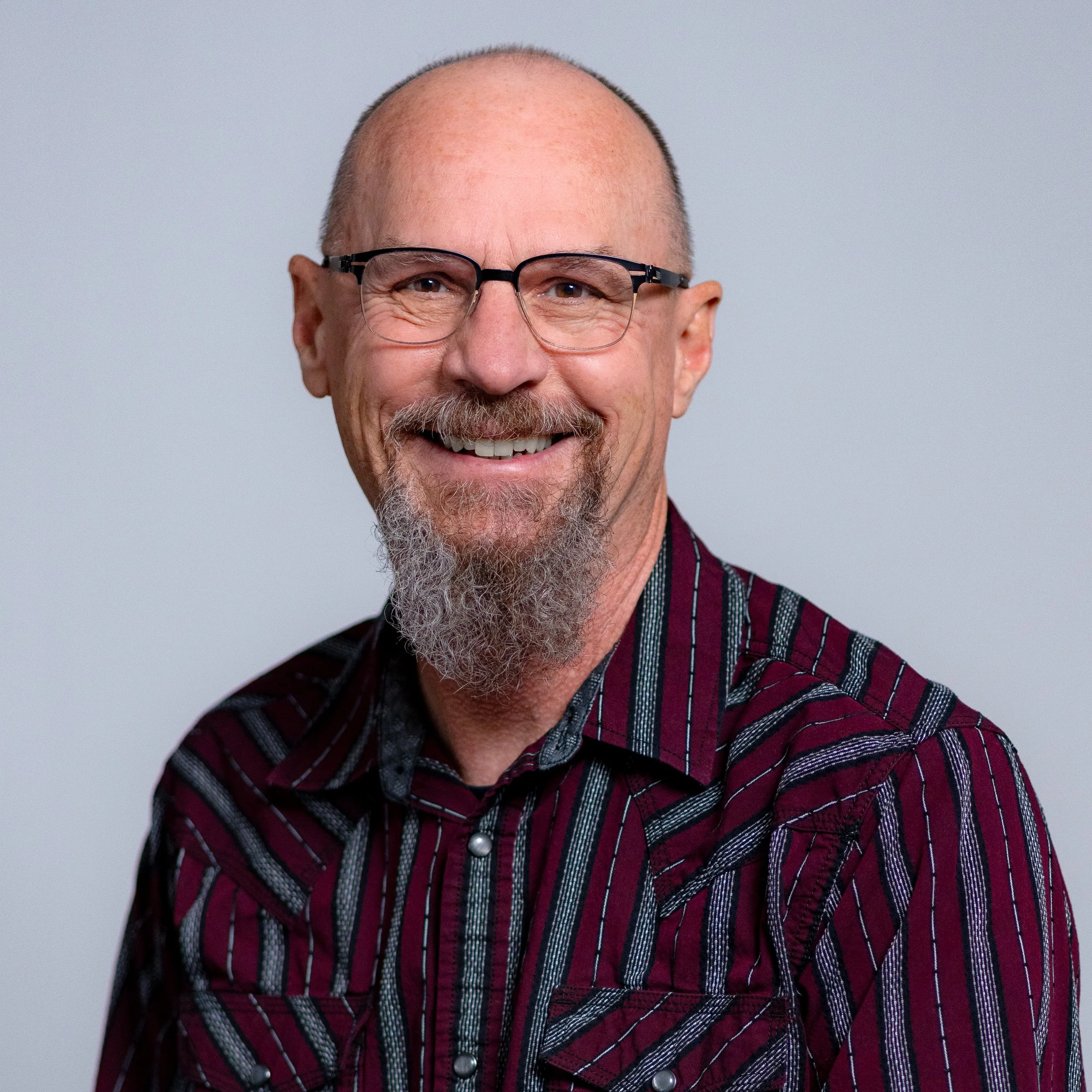 Portrait of a smiling man with glasses, a beard and a goatee, wearing a striped maroon and black shirt, against a plain gray background.