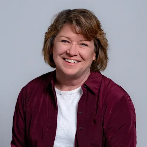 A woman with short, wavy brown hair smiling at the camera, wearing a white t-shirt and a burgundy shirt, against a gray background.