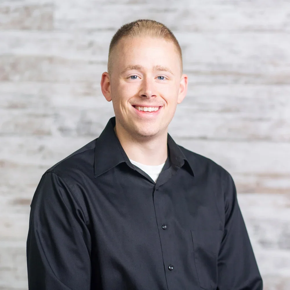 A smiling young man with short light hair, wearing a black button-up shirt, standing against a white textured background.