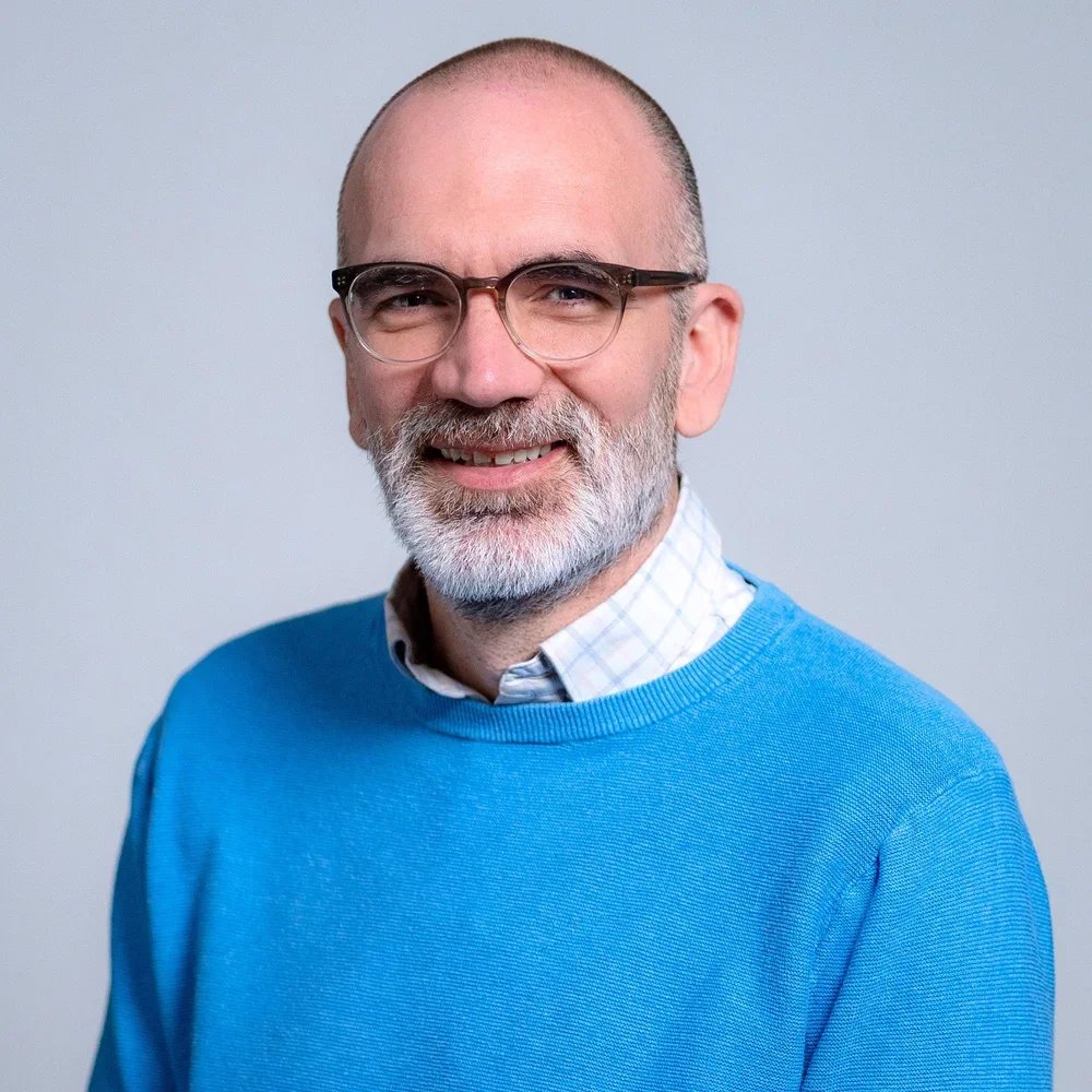 A middle-aged man with glasses, a beard, and a bald head, smiling, wearing a blue sweater over a collared shirt, against a plain light gray background.