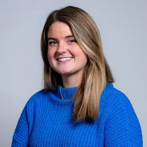A young woman with shoulder-length brown hair, wearing a bright blue sweater, smiling at the camera against a light gray background.