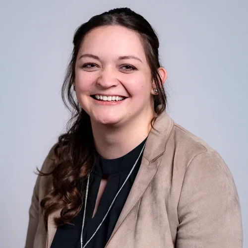 A smiling young woman with long, wavy brown hair, wearing a light brown blazer over a black top, against a light gray background.
