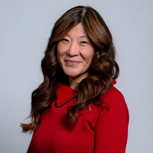A woman with long, wavy brown hair wearing a red top and necklace, smiling at the camera against a plain light background.