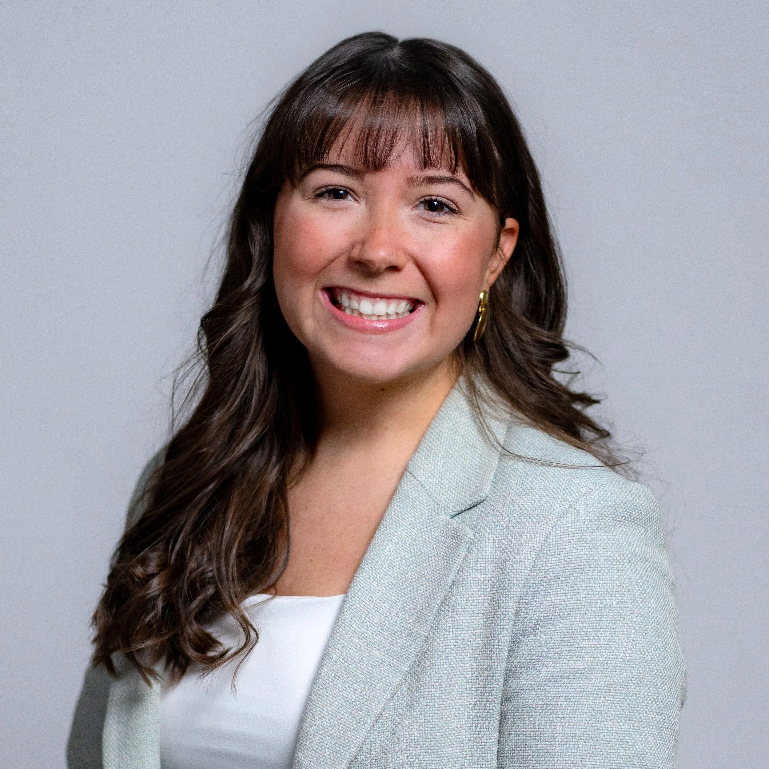 Smiling woman with long dark hair, wearing a light-colored blazer and white top, posing against a plain gray background.