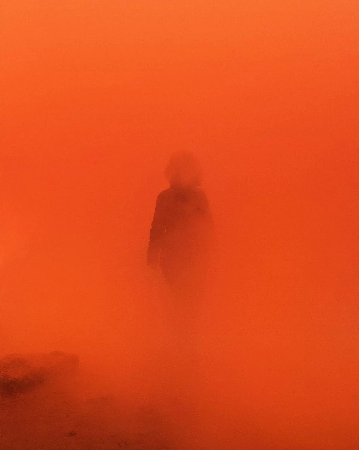 Persona caminando en un entorno cubierto de humo o polvo de color naranja intenso.