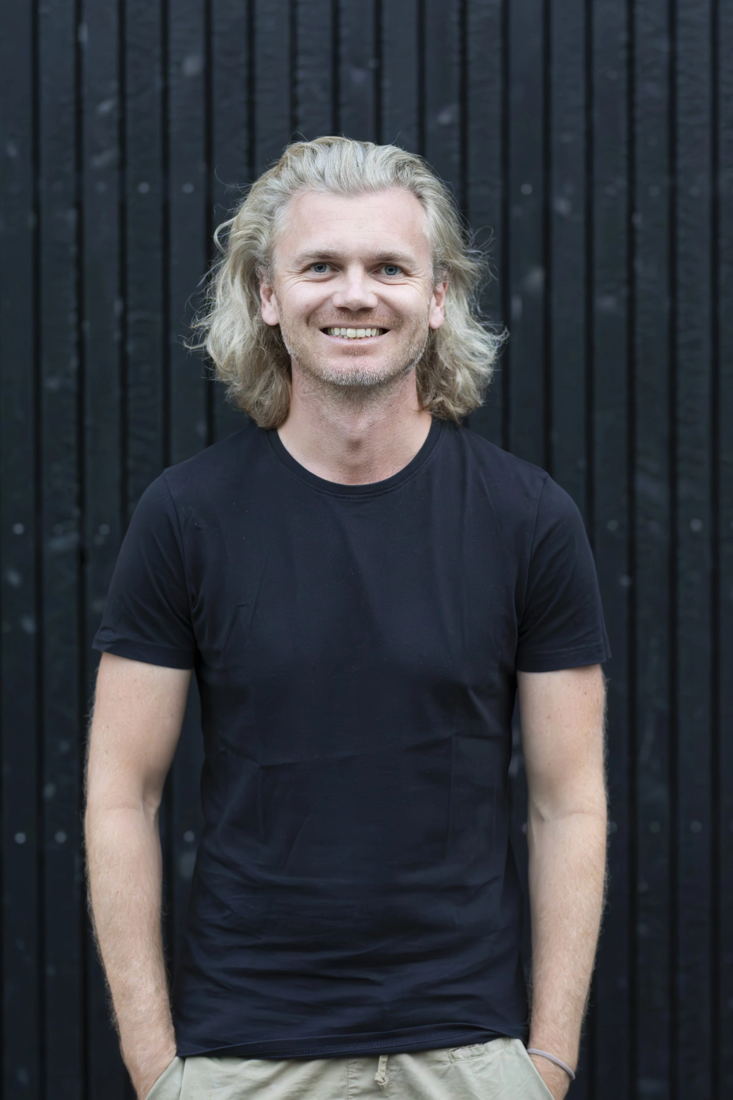 Ein Mann mit langen, lockigen, blonden Haaren, trägt ein schwarzes T-Shirt und steht vor einer schwarzen Holzwand, lächelnd.