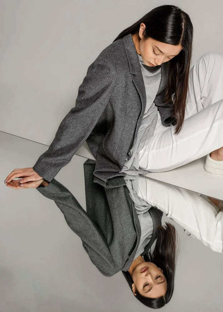 A woman in a gray suit sitting on the floor, with her reflection visible in a shiny surface, creating an upside-down image of her.