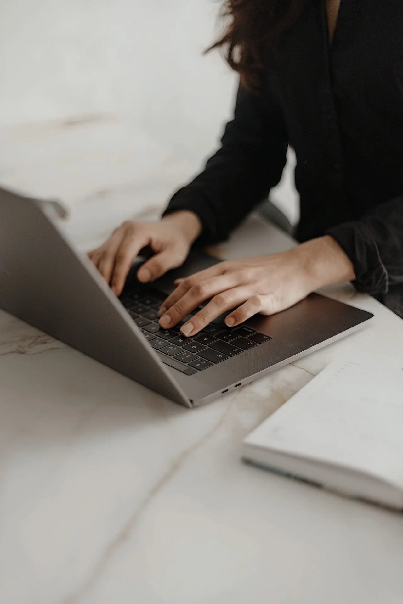 Person wearing a black long-sleeve shirt typing on a silver laptop on a white marble table, with a closed notebook nearby.