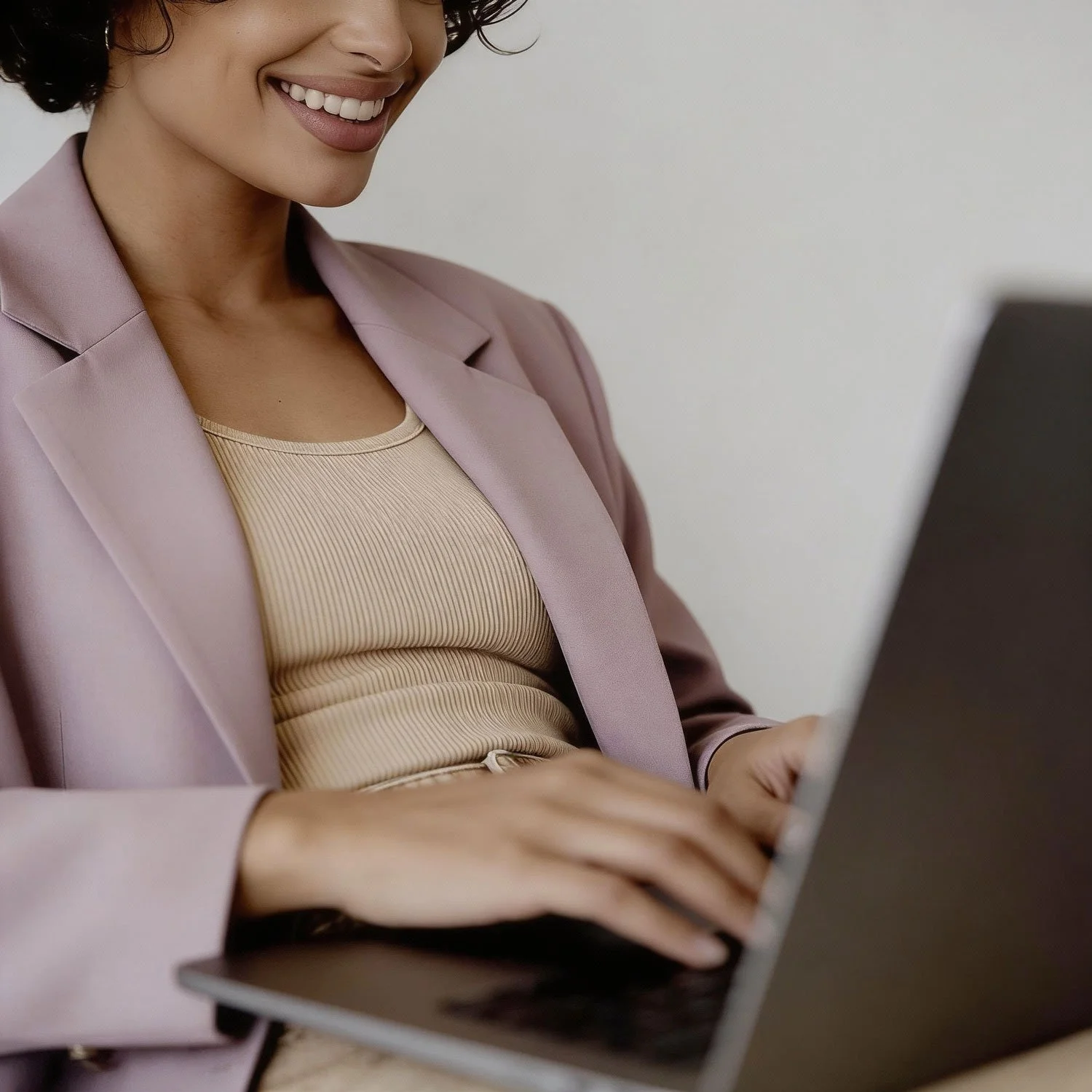 A woman smiling and working on a laptop, wearing a pink blazer and a beige top.