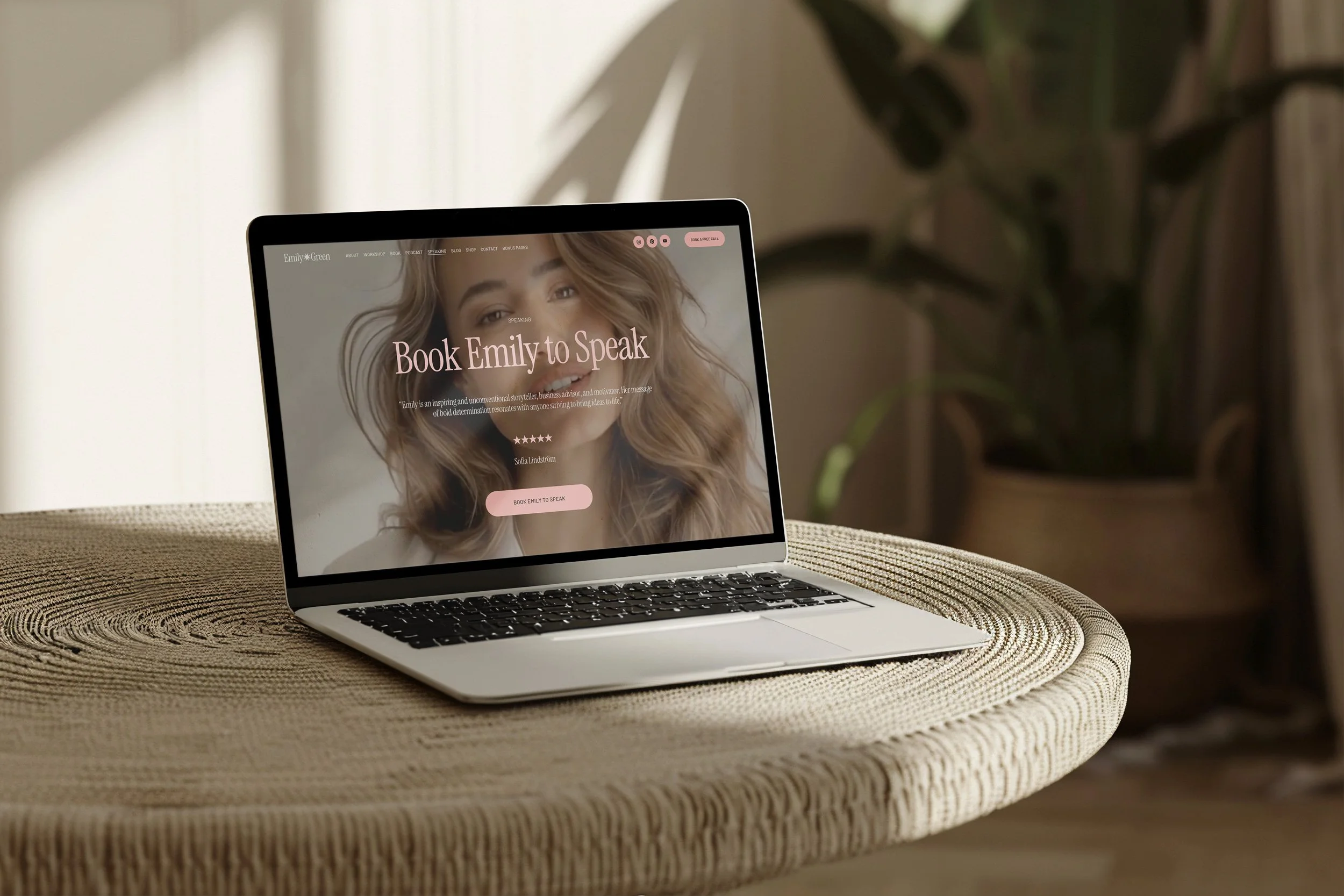 Laptop on a round woven table displaying a website for booking Emily to speak, with a woman's face and pink accents on the screen, in a room with a plant and natural light.
