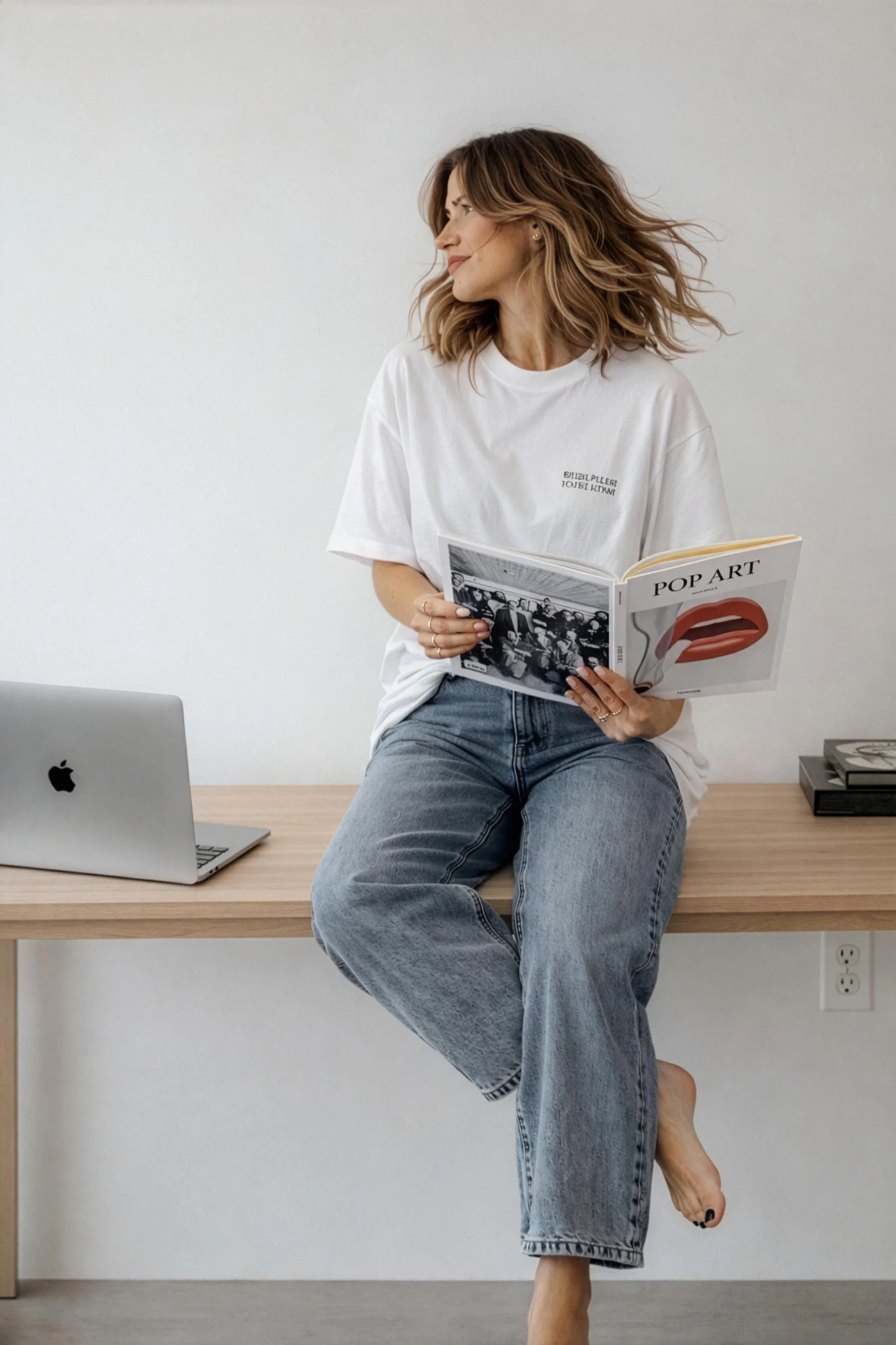 A woman with shoulder-length wavy brown hair, sitting on a wooden desk, holding an art book titled "POP ART." She is wearing a white T-shirt and loose blue jeans, with one foot resting on the desk and the other hanging down. There is a silver Apple MacBook open on the desk and a small stack of books to the side. The background is a plain white wall with an electrical outlet visible.