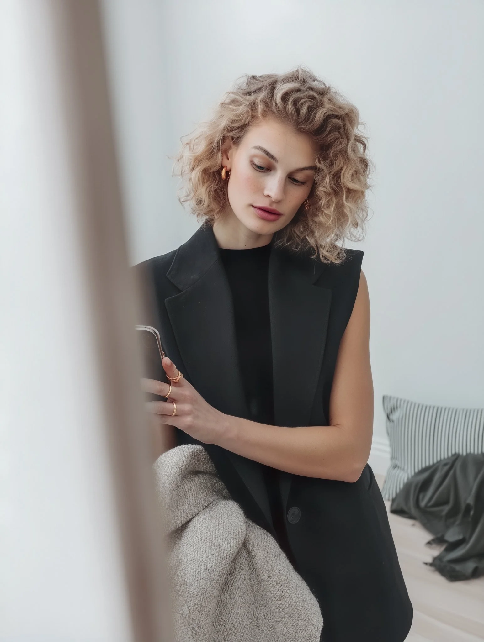 A woman with curly blonde hair, wearing a black sleeveless blazer, looking at her phone, standing in a modern, minimalist room with neutral colors.
