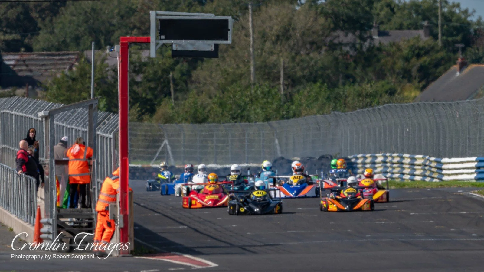A group of go-kart race cars on a racing track, with race officials and spectators on the side