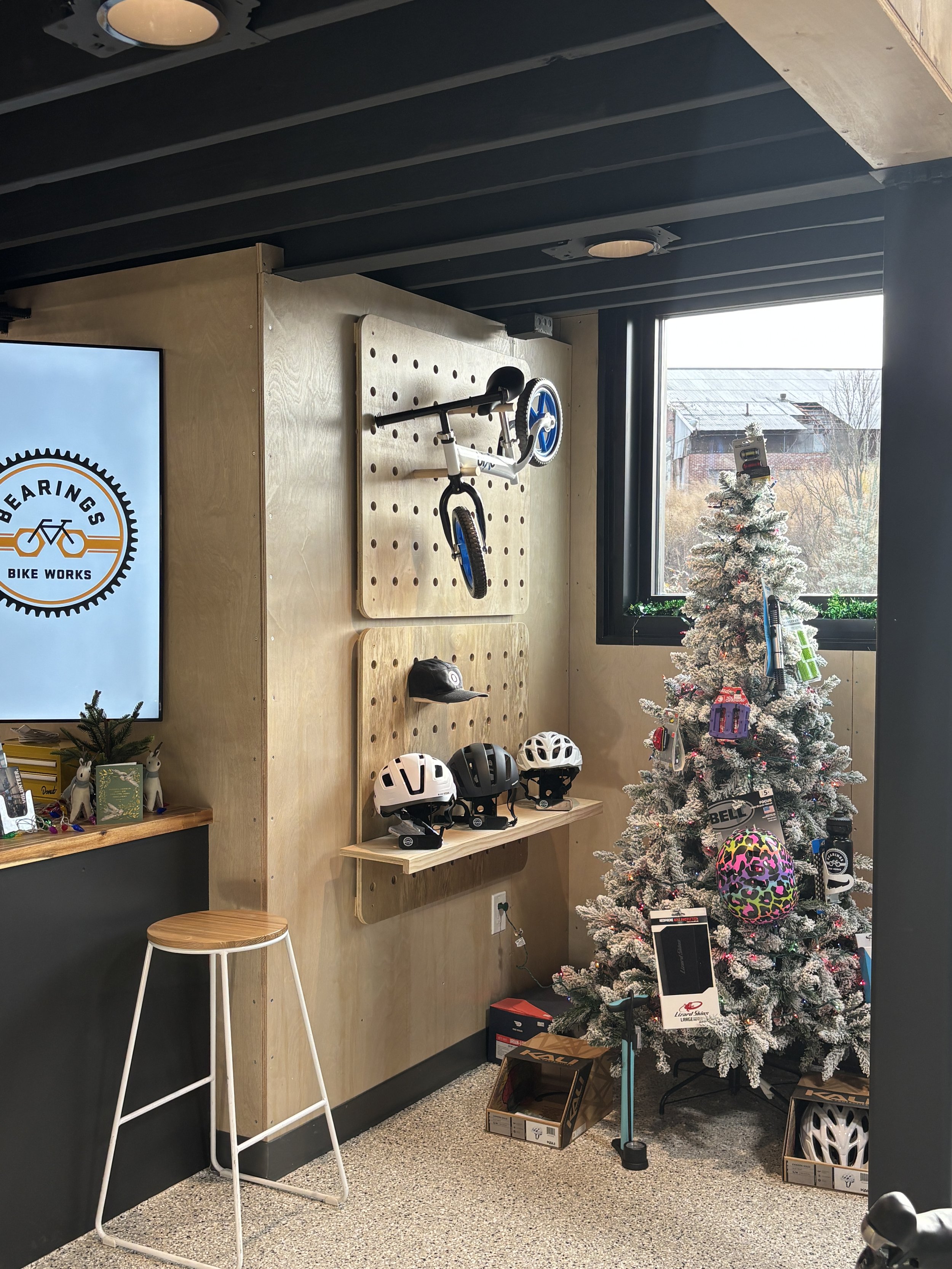 Indoor bike shop with a Christmas tree decorated with bike accessories, helmets, and a scooter mounted on a wooden pegboard, next to a window showing outdoor scenery.