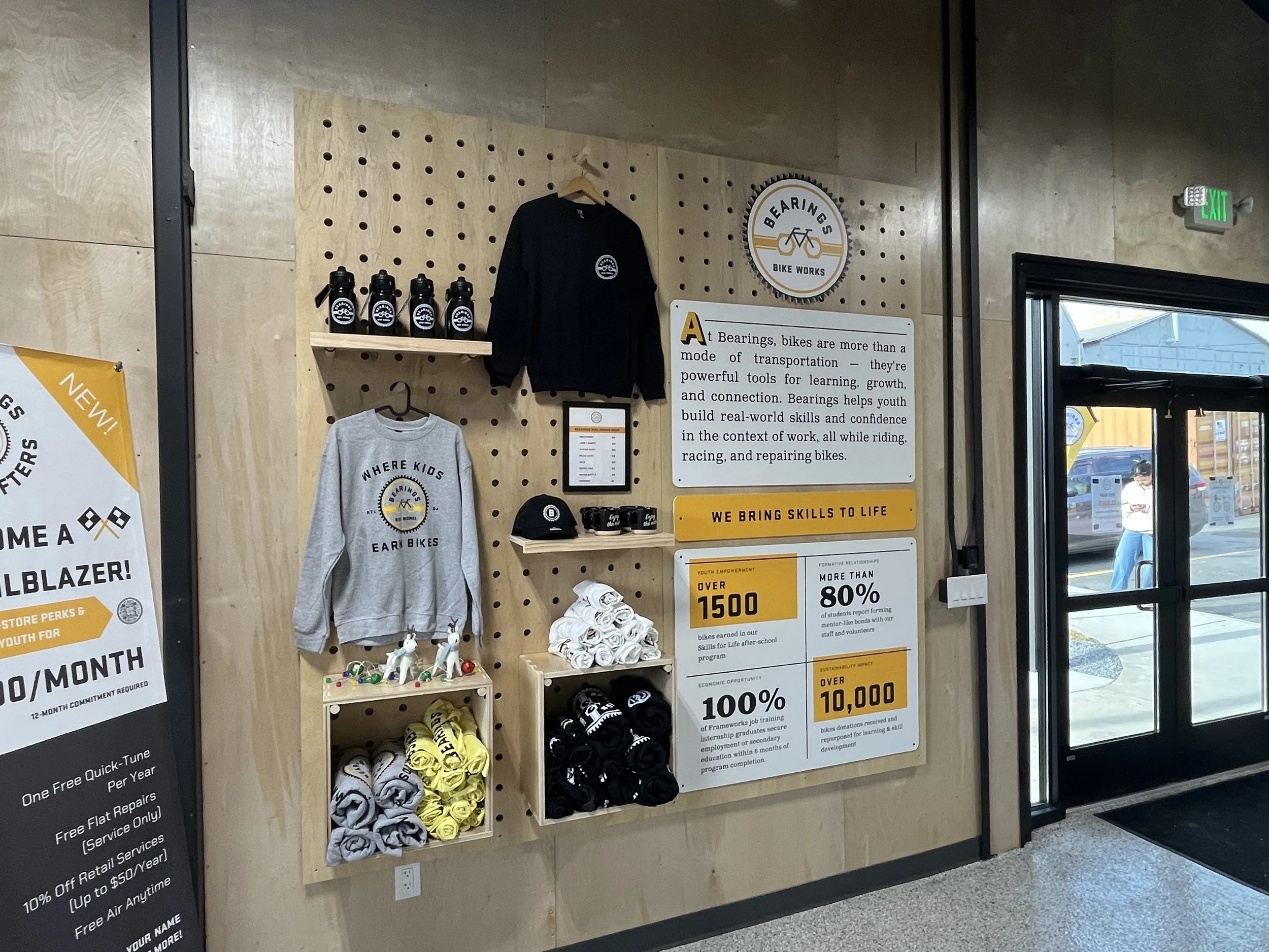 Interior of a bike shop with a display wall featuring branded water bottles, clothing, caps, and rolled-up towels, alongside informational signs about the shop's programs and achievements.