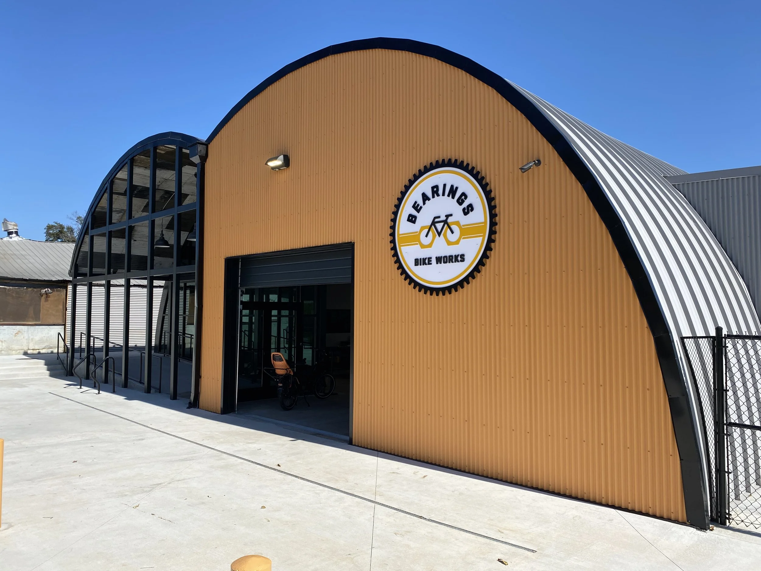A modern building with a rounded roof and large windows, labeled 'Bearings Bike Works,' with a yellow and black logo featuring a bicycle design, under a clear blue sky.