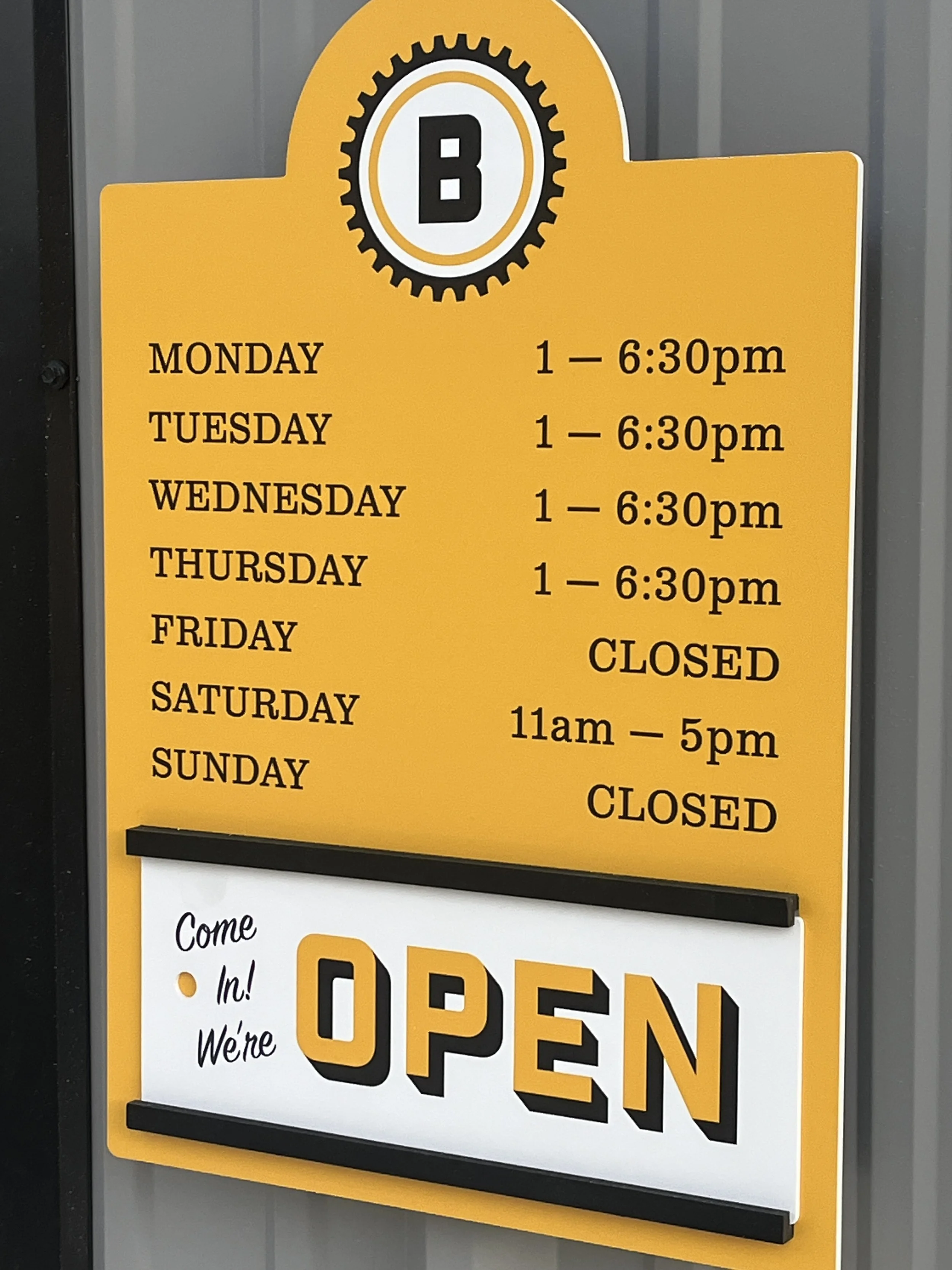 Business hours sign with a yellow background, black text, and a large sign reading "Come in! We're OPEN" in front. The sign lists weekday hours from 1 pm to 6:30 pm (Monday through Thursday), closed Fridays, Saturday hours from 11 am to 5 pm, and closed Sundays.