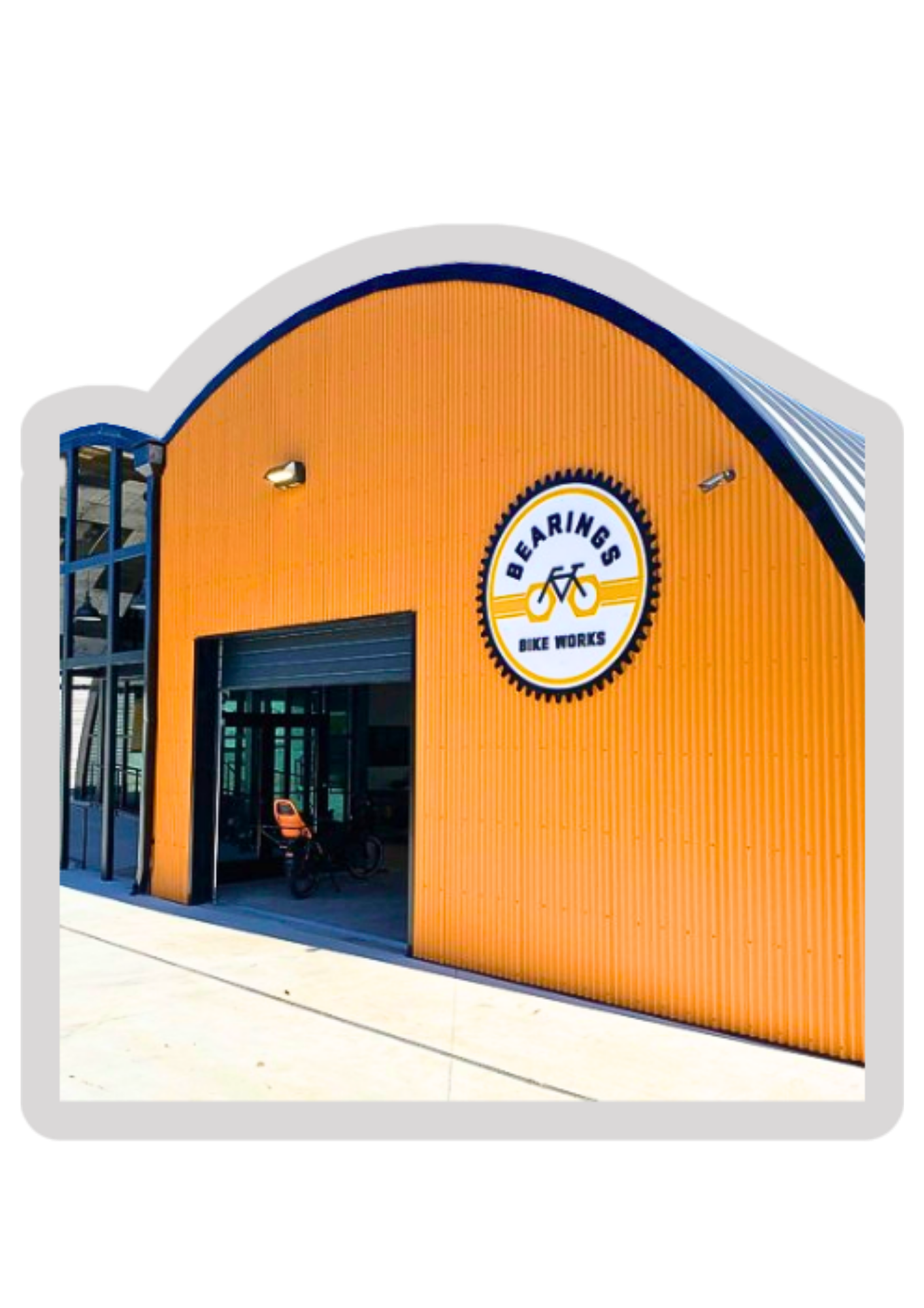 Exterior view of a bike shop named Bearings Bike Works with a bright orange corrugated metal facade and a large logo featuring a bicycle gear and the shop's name.
