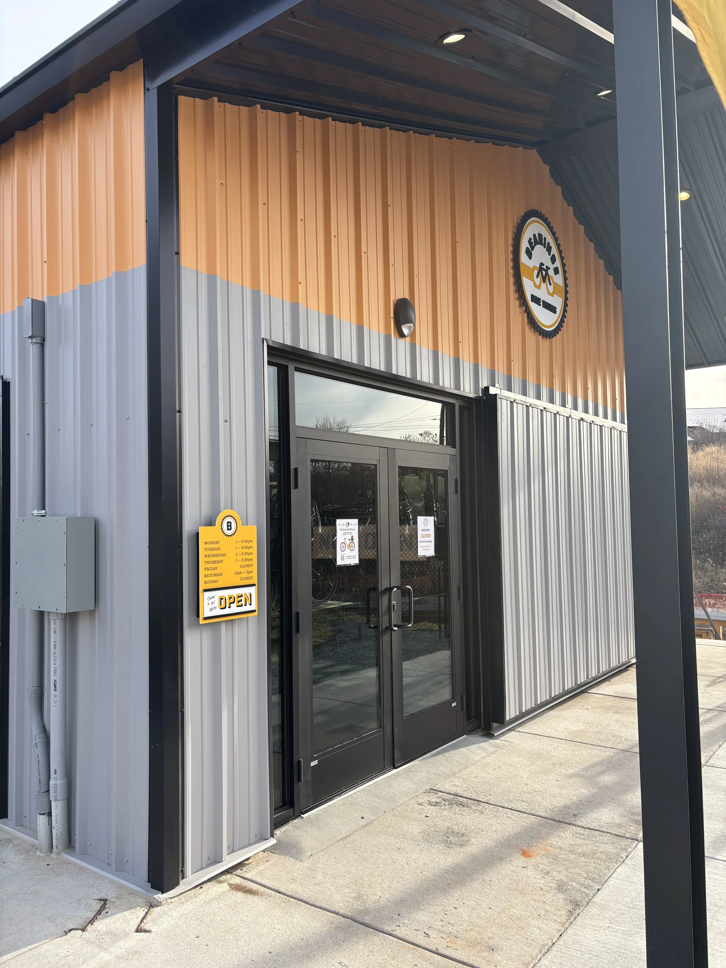 Front entrance of a bike shop with a black glass door, orange and gray metal siding, a small sign reading 'OPEN', and a logo with a bike and gears above the door.