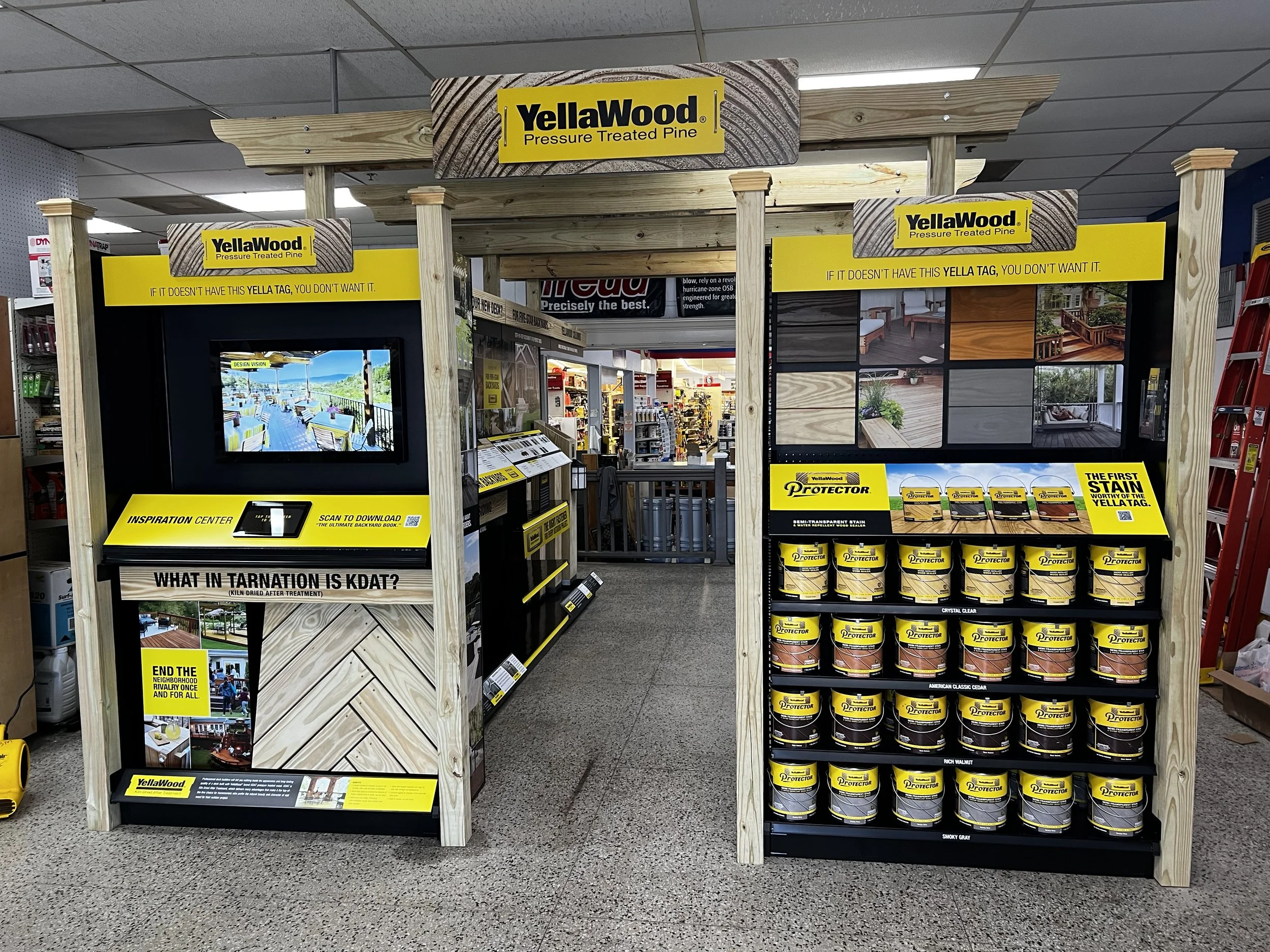Display of pressure-treated pine wood, YellaWood brand, in a hardware store aisle with yellow signage and cans of wood stain, showing samples of wood finishes and projects.