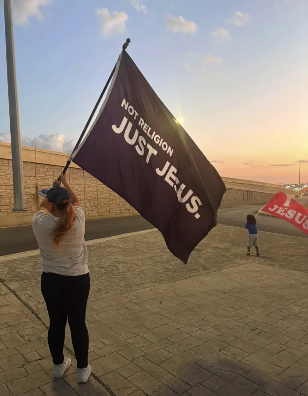 A woman holding a large black flag that reads 'Not Religion Just Jesus' stands on a paved sidewalk near a stone wall as the sun sets. A child in the background holds a red flag.