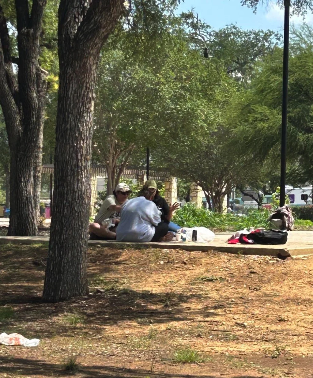 People sitting on a park bench under trees, engaging with their phones, with backpacks and water bottles nearby, on a sunny day.