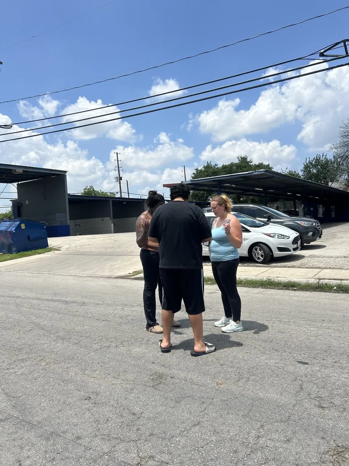 Three people standing in a parking lot, looking at a phone together, with cars and a building with a covered parking area in the background.