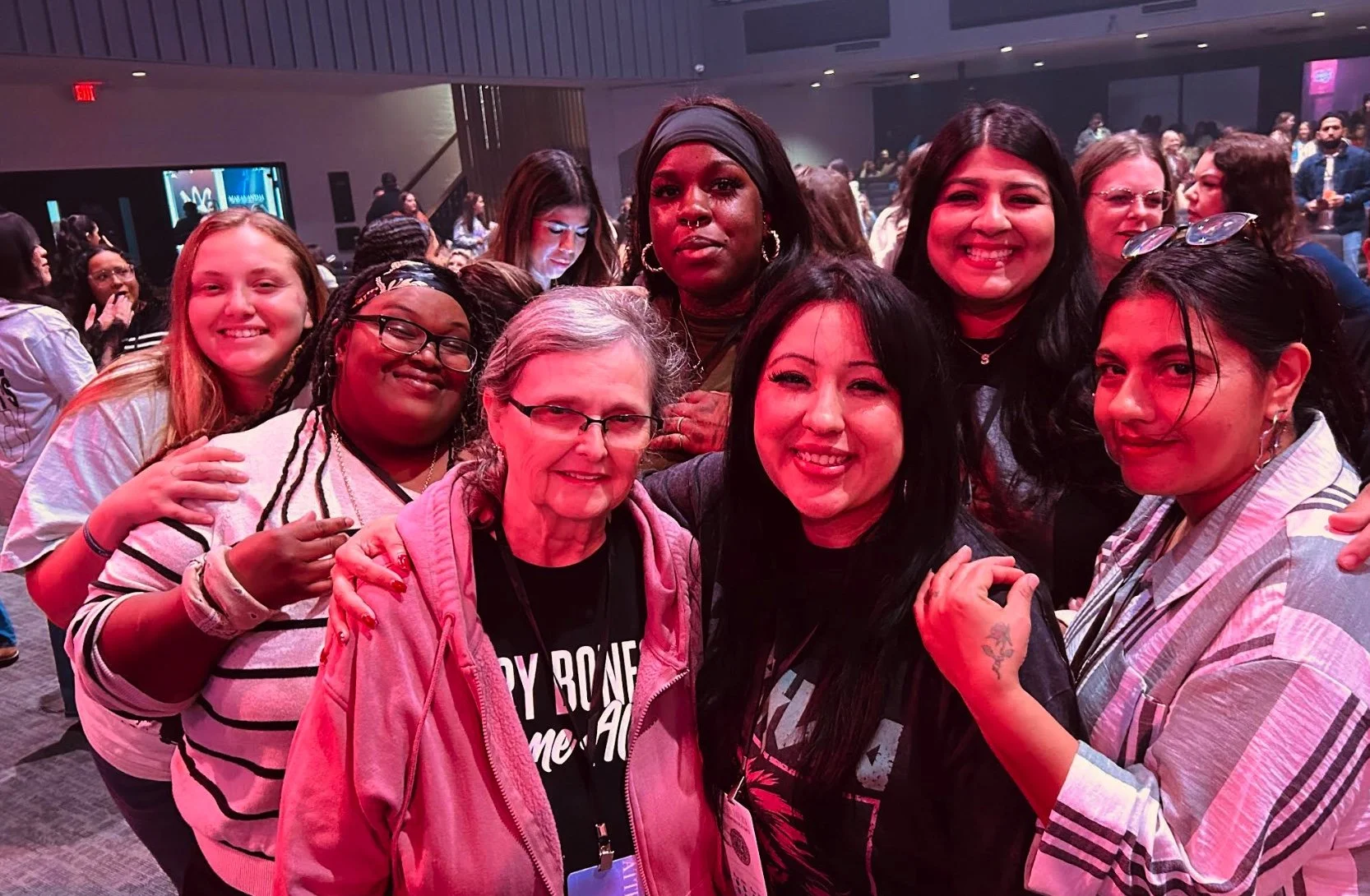 Group of diverse women smiling together at an indoor event, with others in the background.