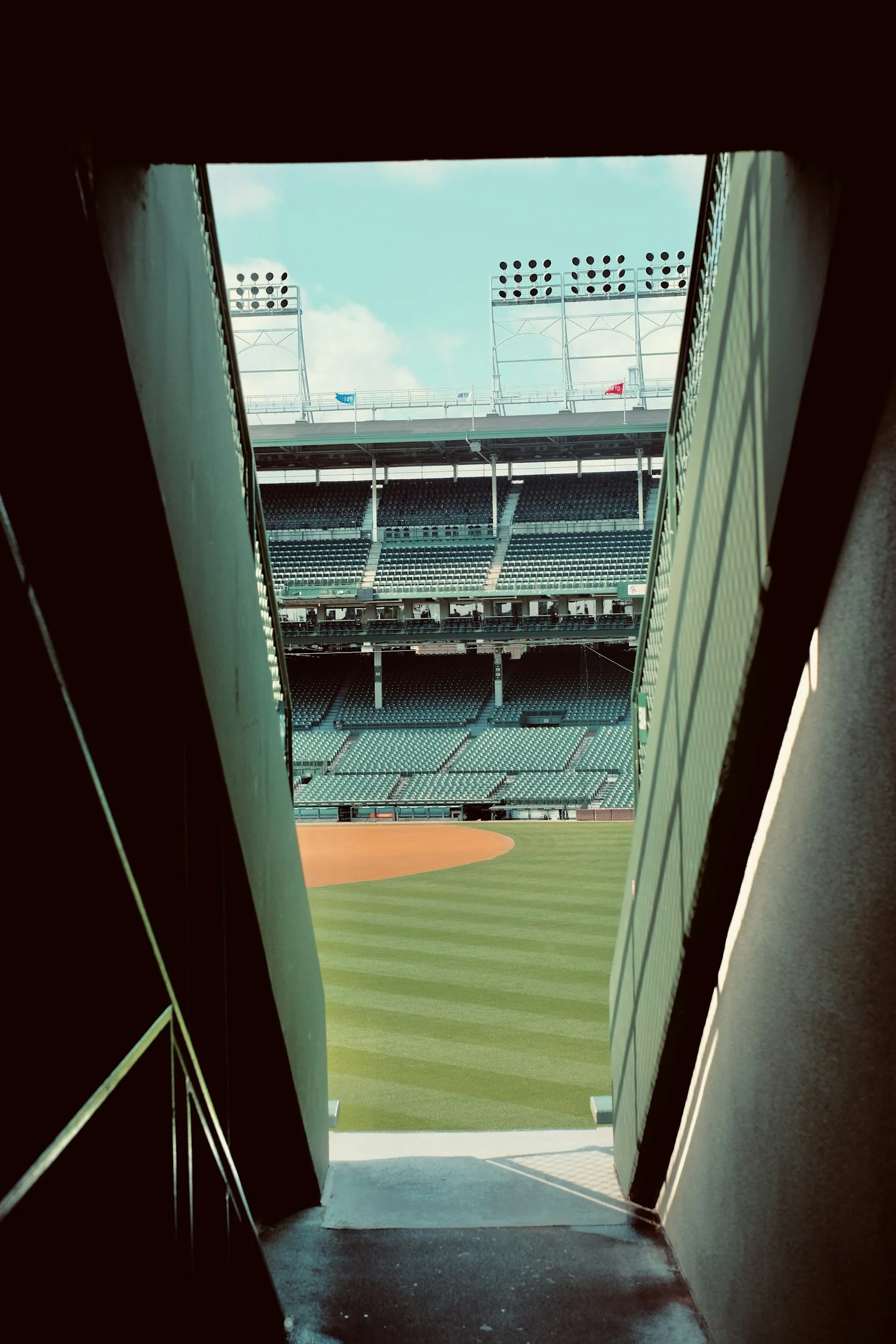 View of a baseball stadium through a narrow opening, showing green seating and a field with a dirt infield and green grass outfield.