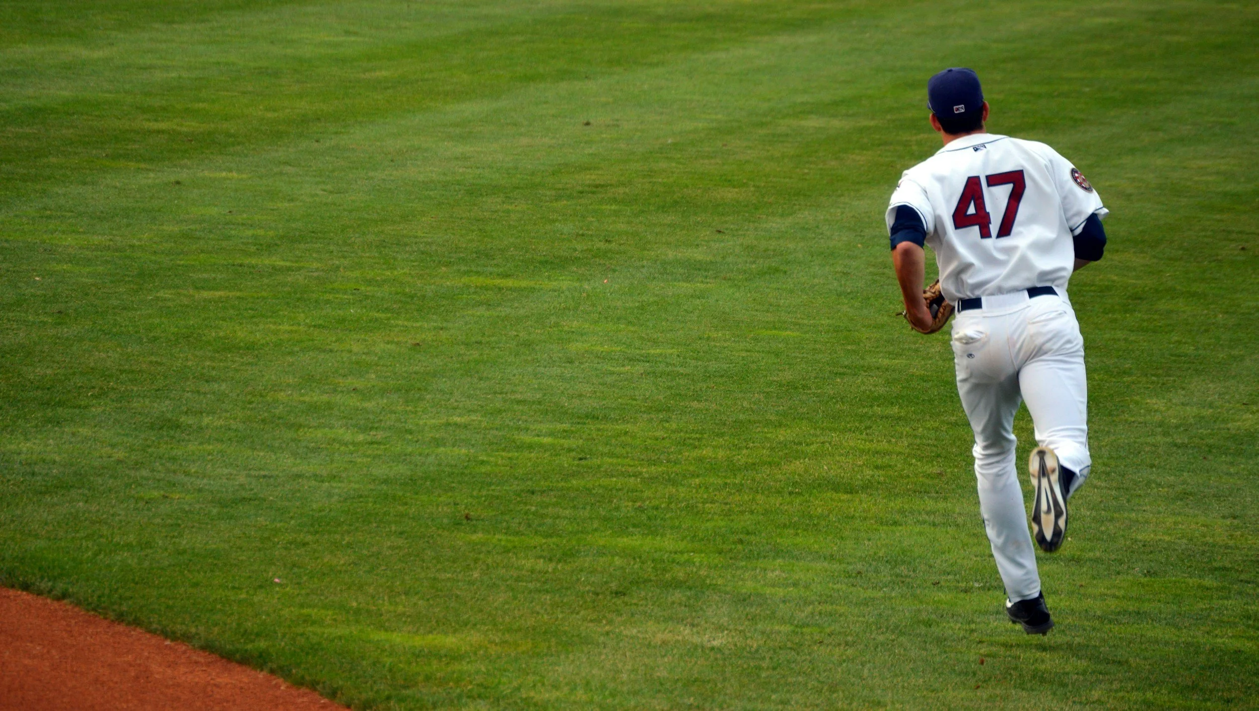 A baseball player wearing a white uniform with the number 47 on the back, running on the grassy field with green grass and a dirt edge in the baseball outfield.