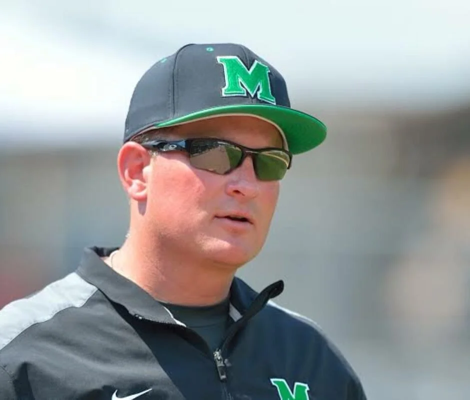 A man wearing a black cap with a green and white letter M, black sunglasses, and a gray sports jacket with black accents, outdoors in a sunny setting.