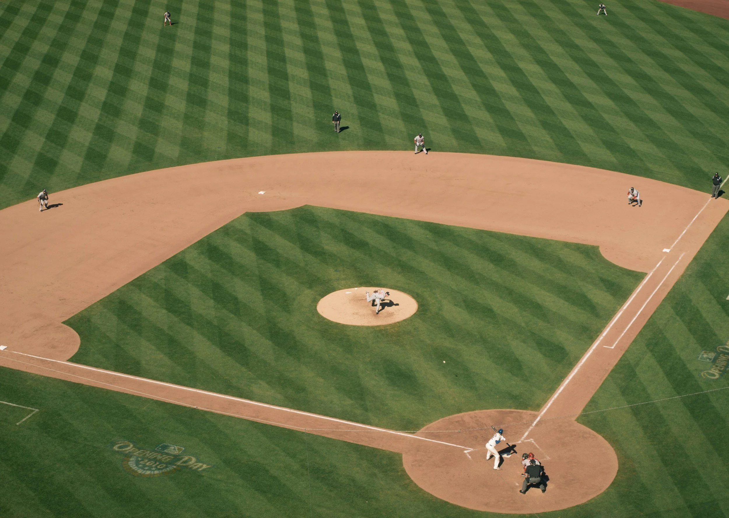 Aerial view of a baseball field with players in position, including the pitcher and catcher, and fielders on the grass and in the outfield.