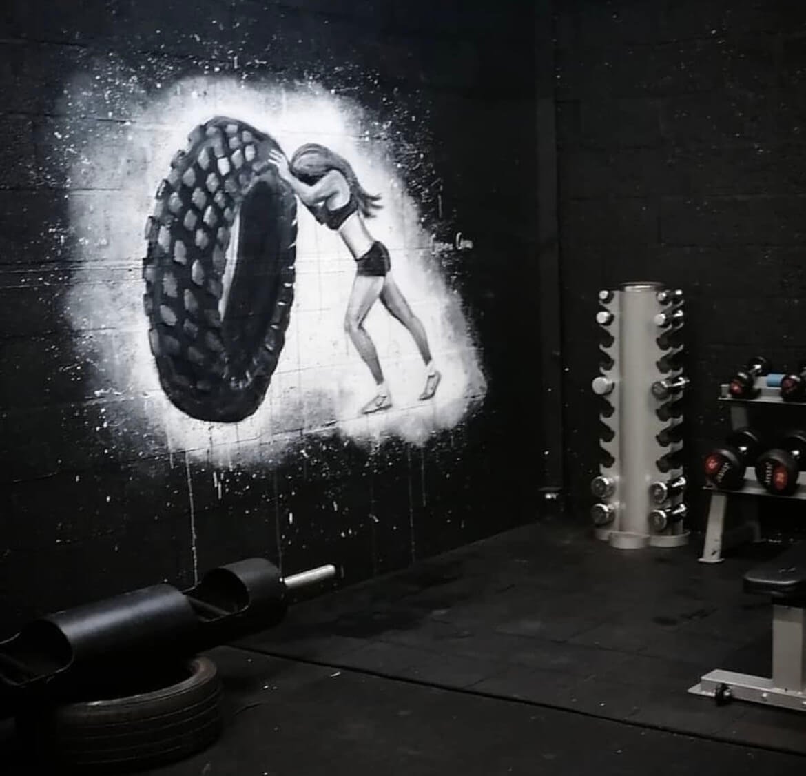 A gym wall mural depicting a girl in fitness gear pushing a large tire, with dumbbells and workout equipment nearby.