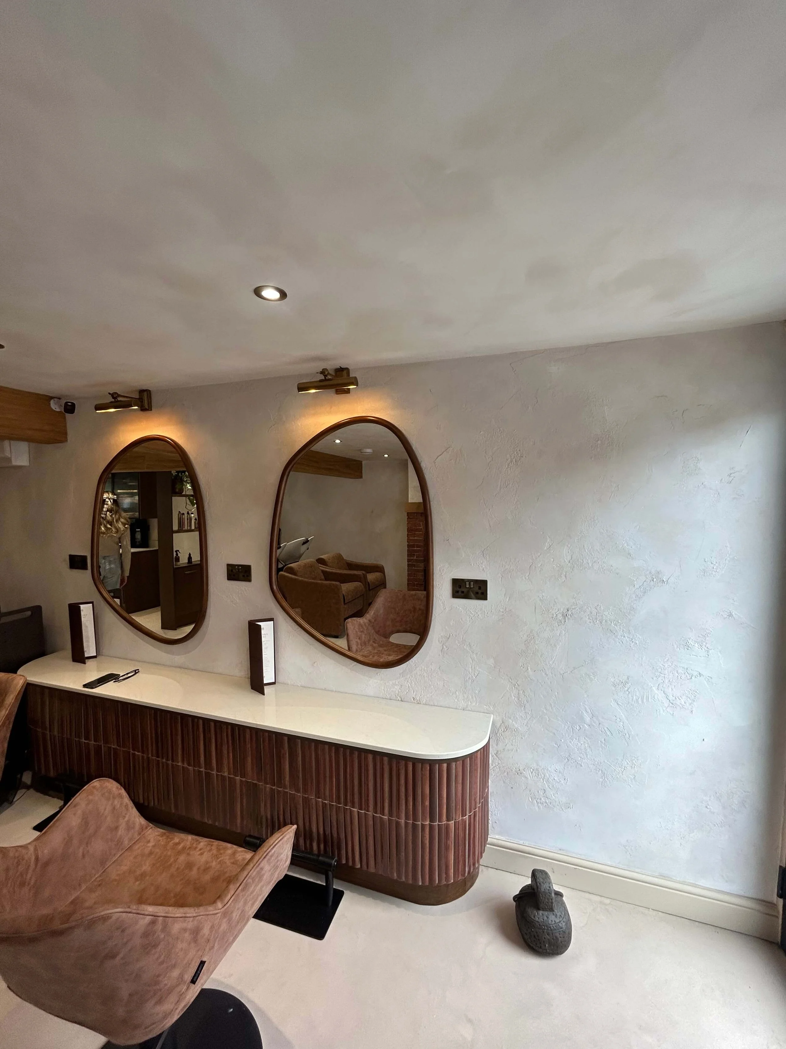 Interior view of a room with two oval mirrors on a textured wall, brown chairs, and a white countertop with a wooden base, with a stone sculpture on the floor near the wall.