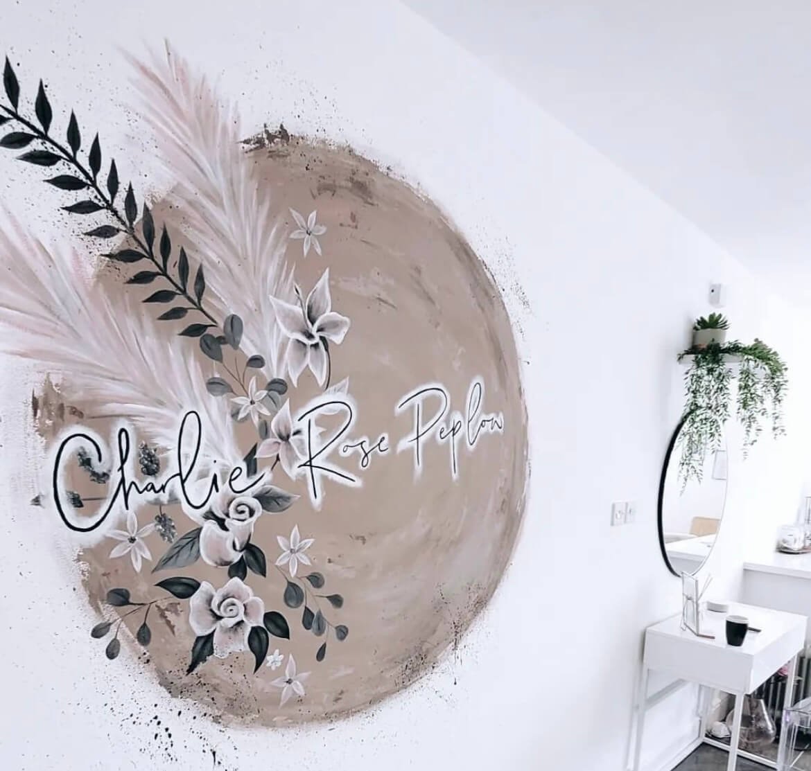 Decorative wall with a floral design and the name Charlie Rose Peplow written in script, next to a mirror and a small table with plants.