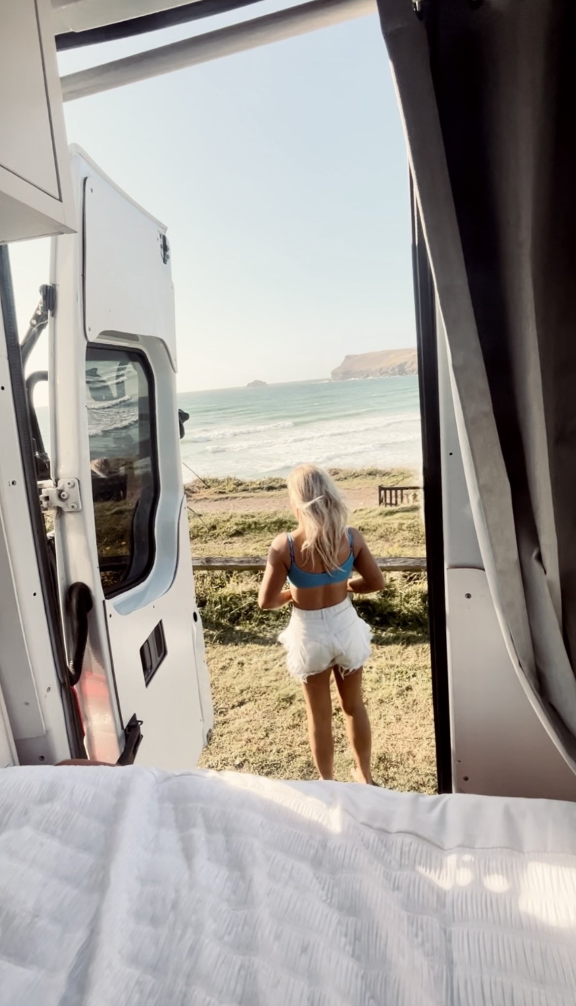View from inside a camper van looking out at a woman in a blue top and white shorts standing on a grassy area near the beach, with ocean waves and a distant land formation in the background.