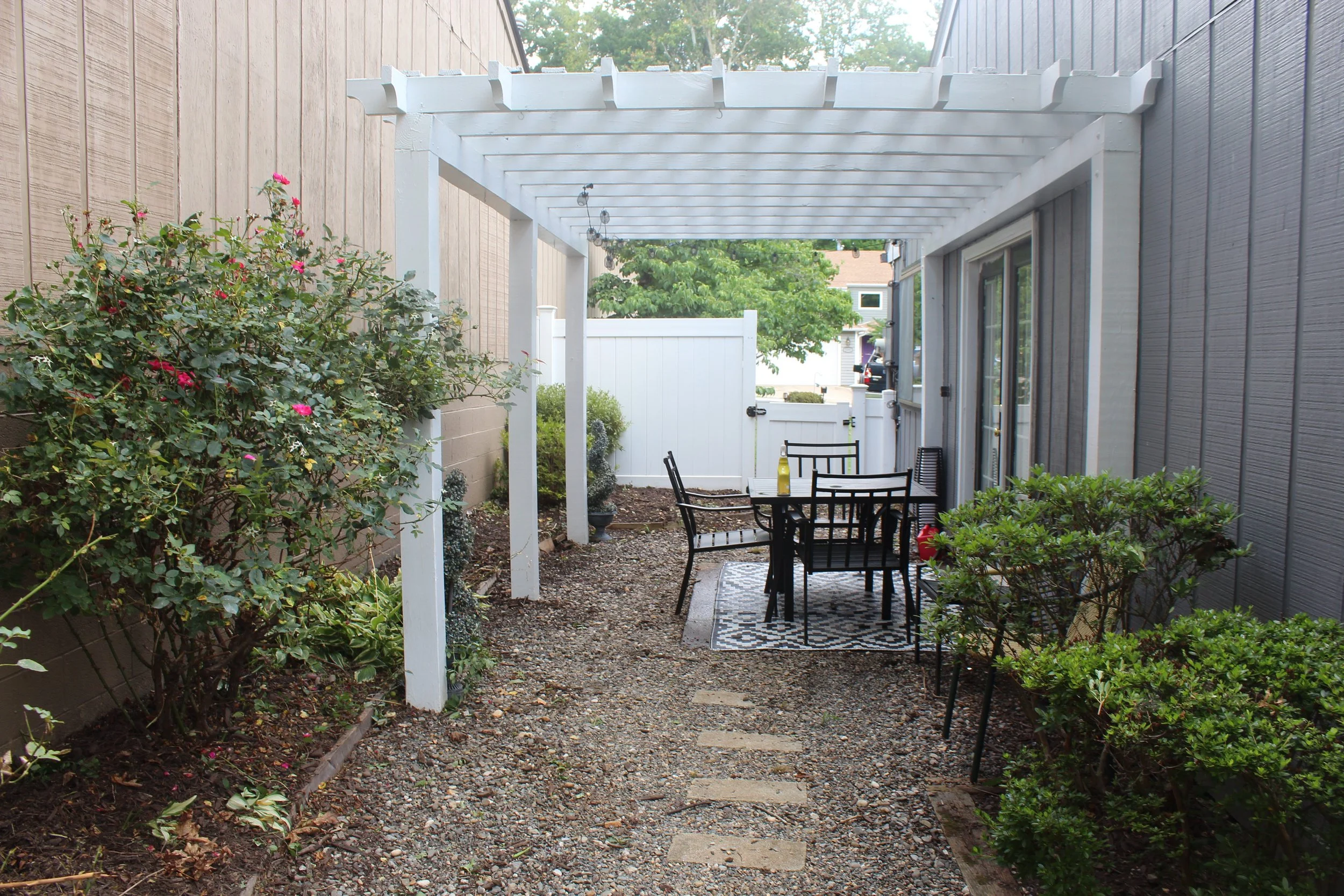 A backyard patio with a white wooden pergola, outdoor table with four chairs, surrounded by bushes and plants, and a gravel pathway leading to a white fence and neighboring houses.