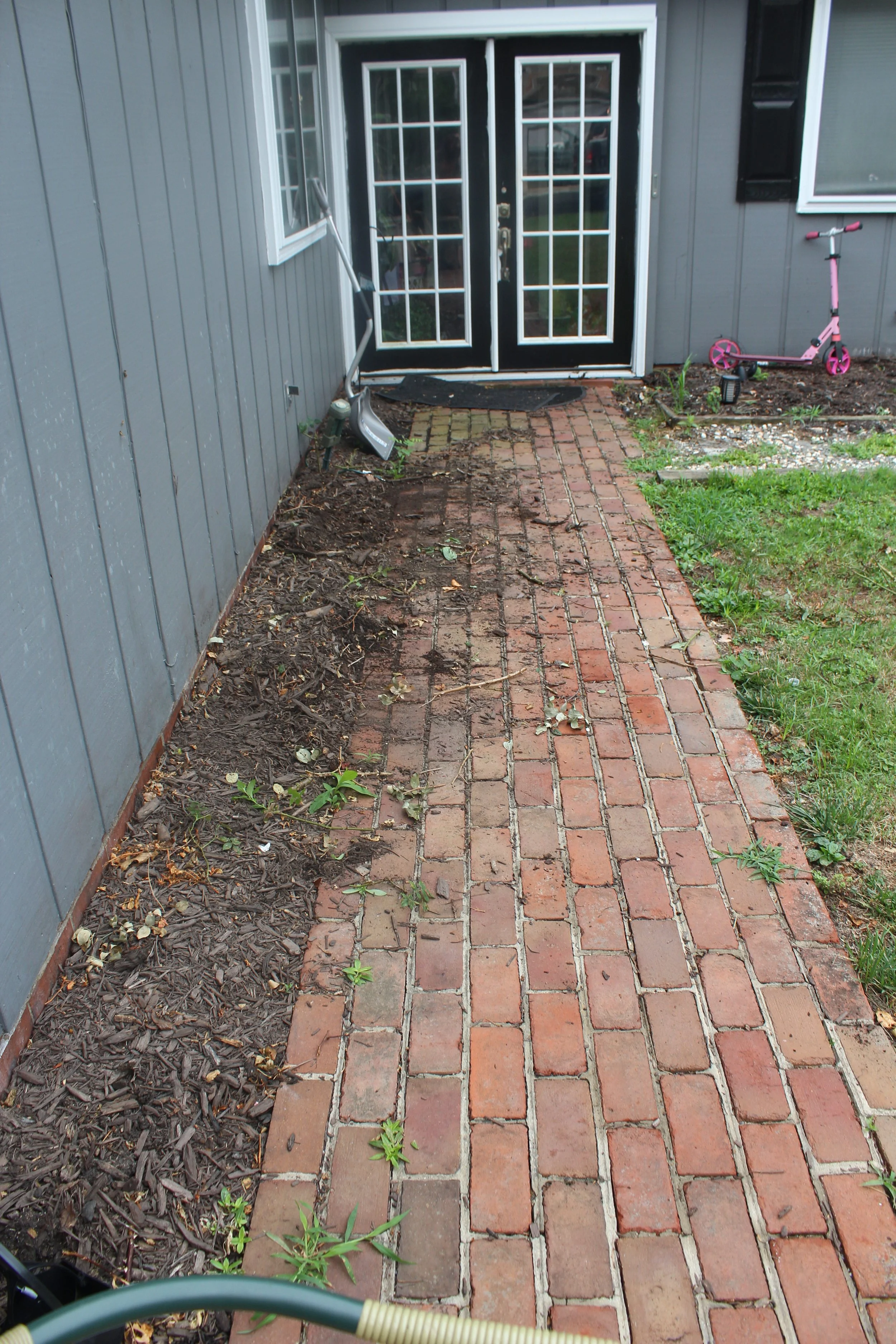 A brick pathway leading to a glass double door entrance of a house, with a gray exterior wall and a window on each side. There are cleaning tools leaning against the wall and a pink child's scooter on the right side. The pathway has some green plants