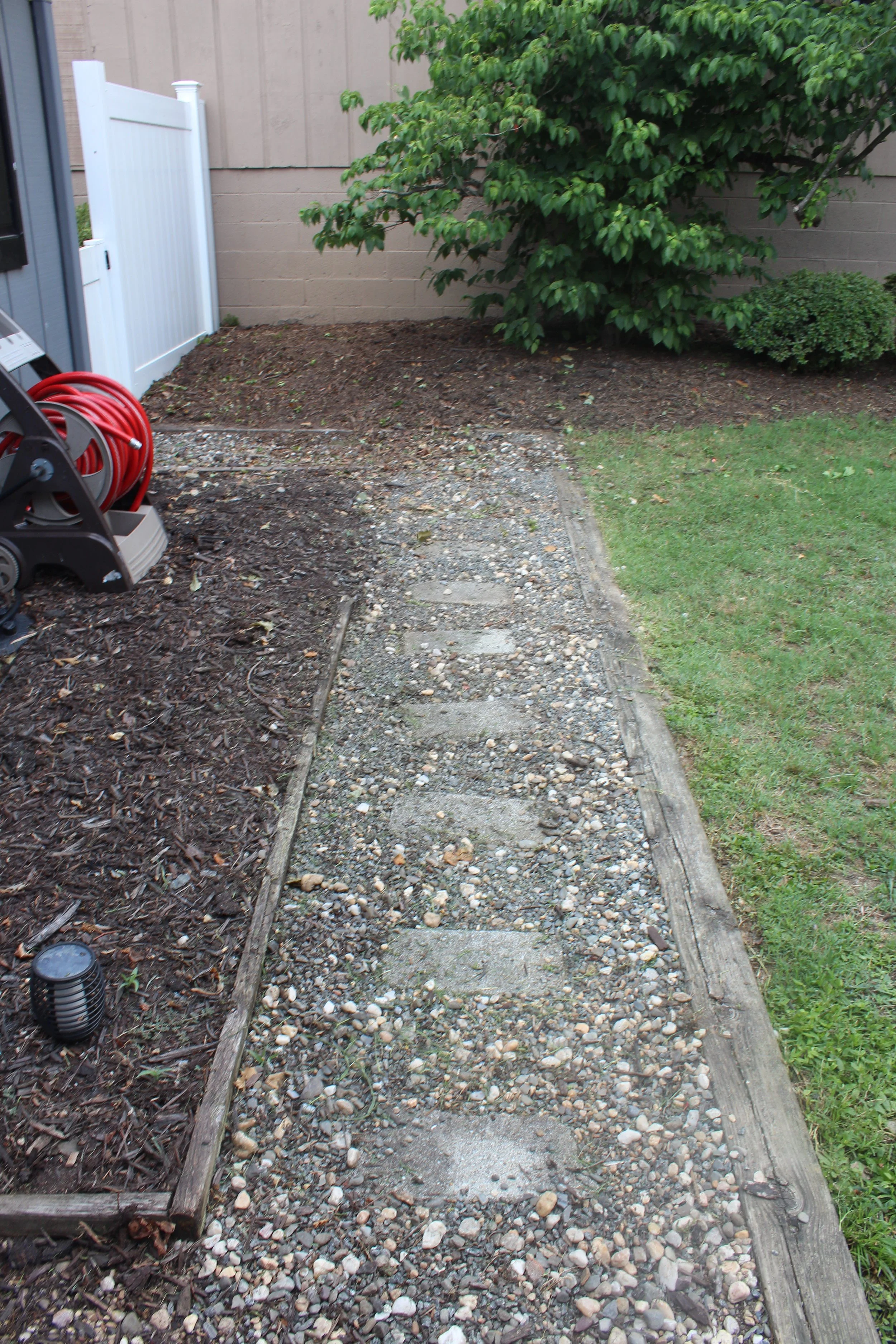 A backyard garden area being prepared for gravel or stone filling, with a partially filled path of small stones, a wooden frame outline, a garden hose reel, and some green bushes against a beige wall.