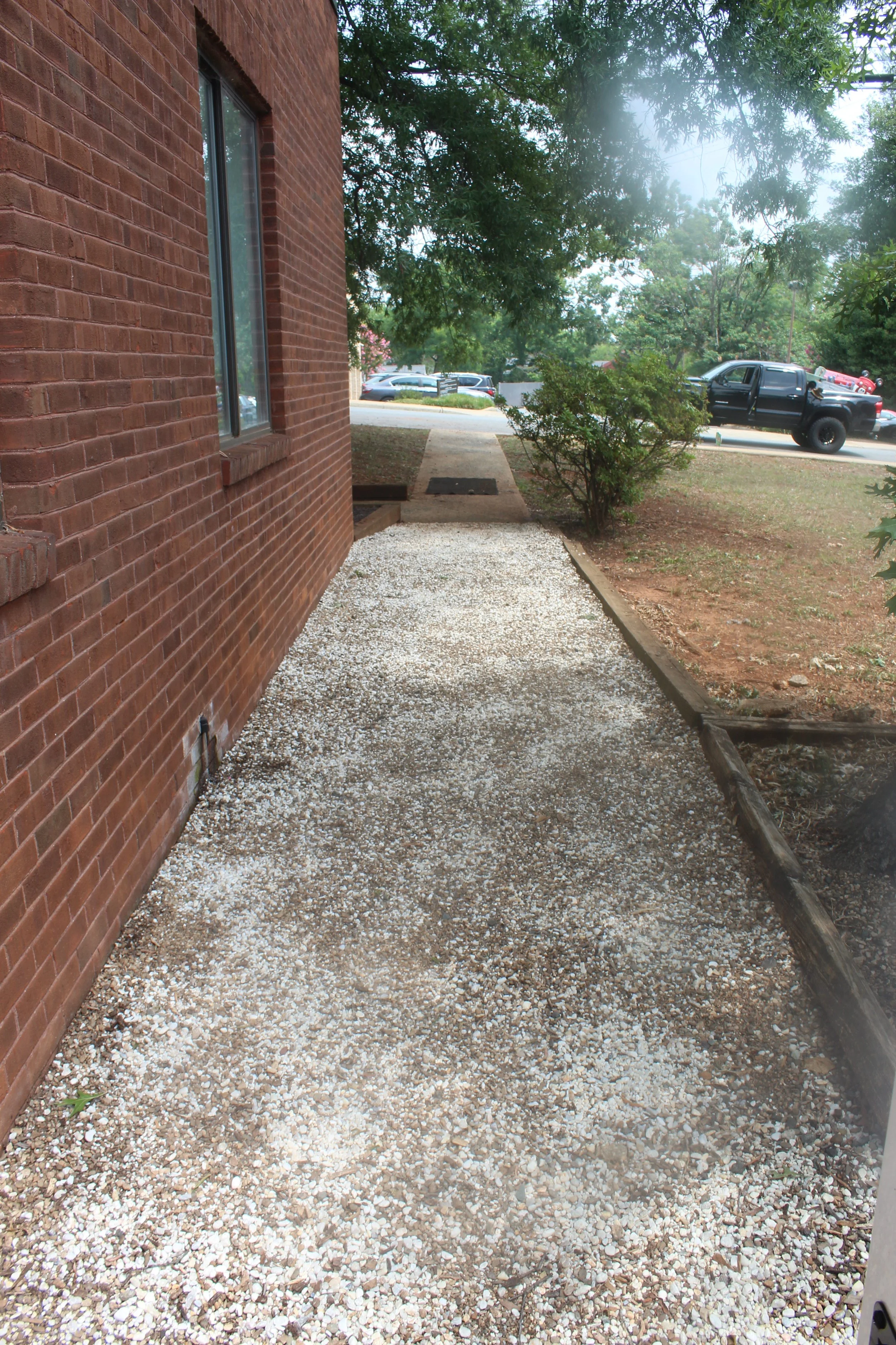 Side view of a brick building with a window, a gravel pathway, and a small shrub. Parked cars are visible in the background along the street, with trees and shrubs also present.