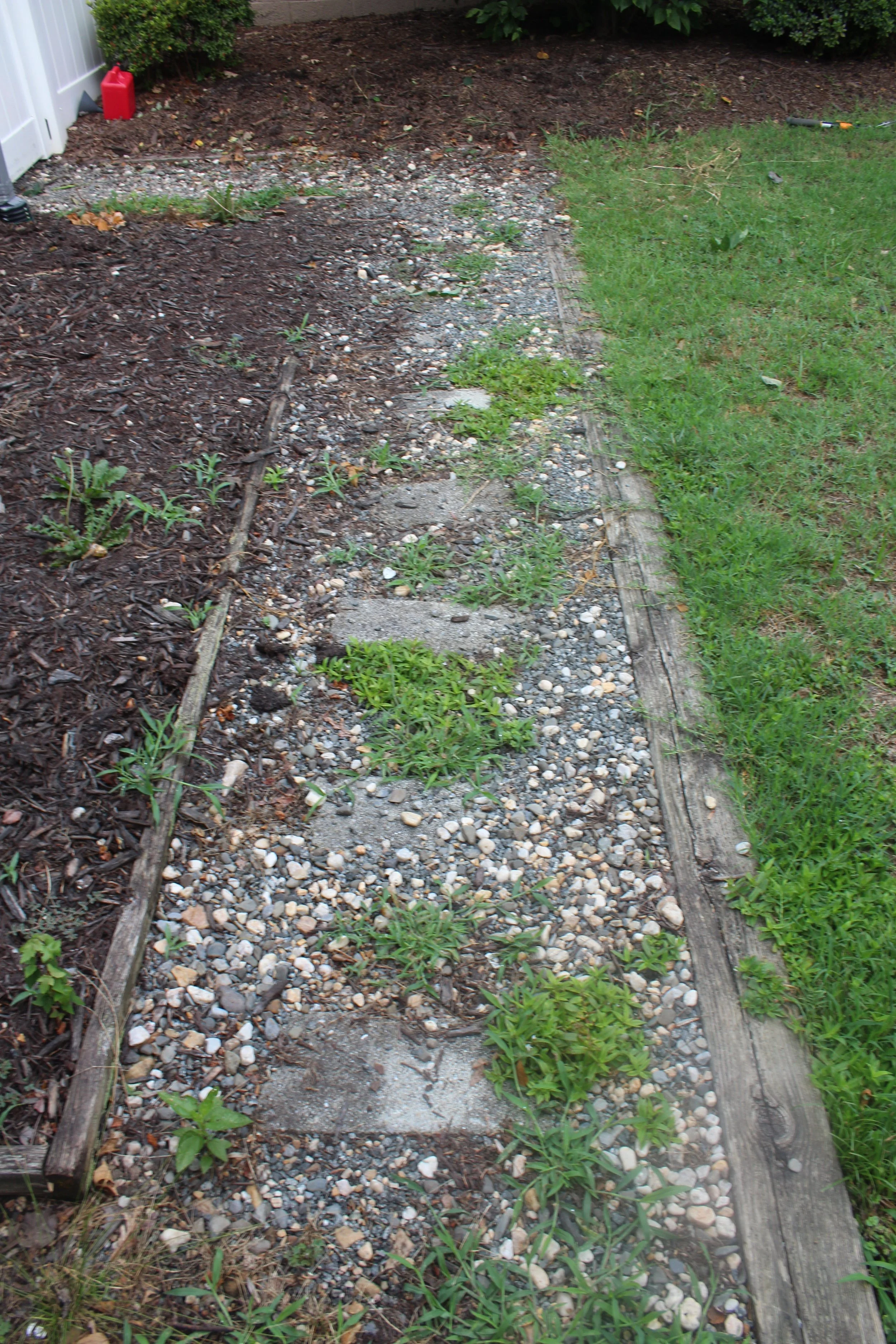 A backyard with a garden bed on the left side filled with soil and small plants, bordered by old wooden planks, and a patch of green grass on the right side.
