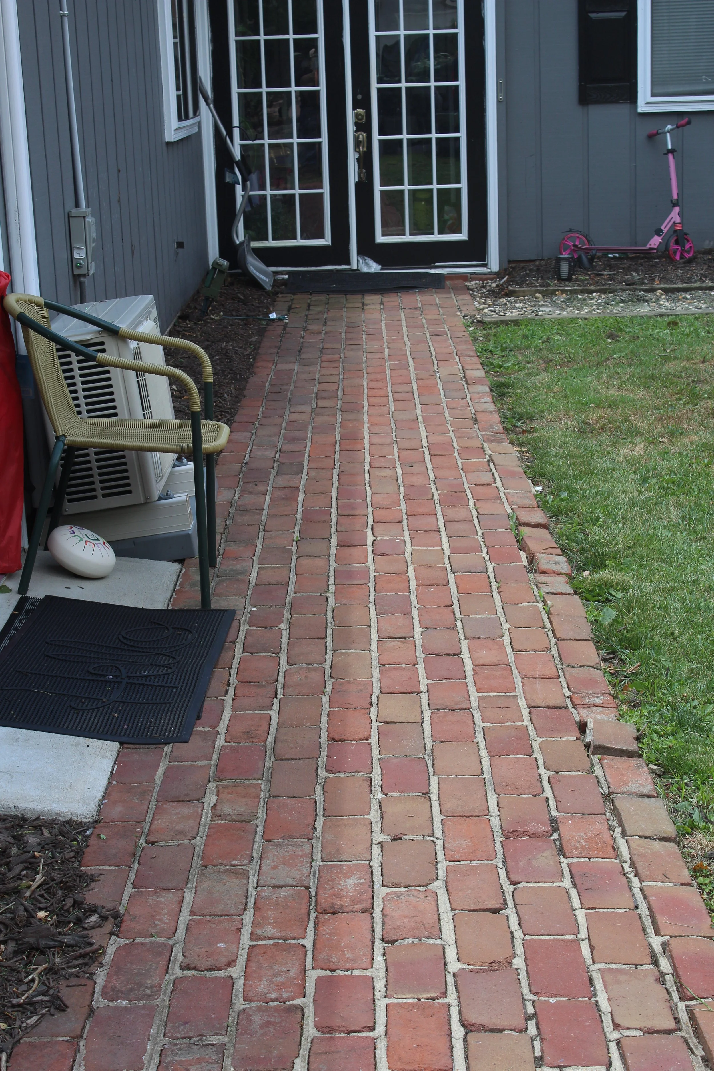 A brick pathway leading to a house with a black door and glass panels, surrounded by a small green lawn and children’s toys, including a pink scooter, on the right side.