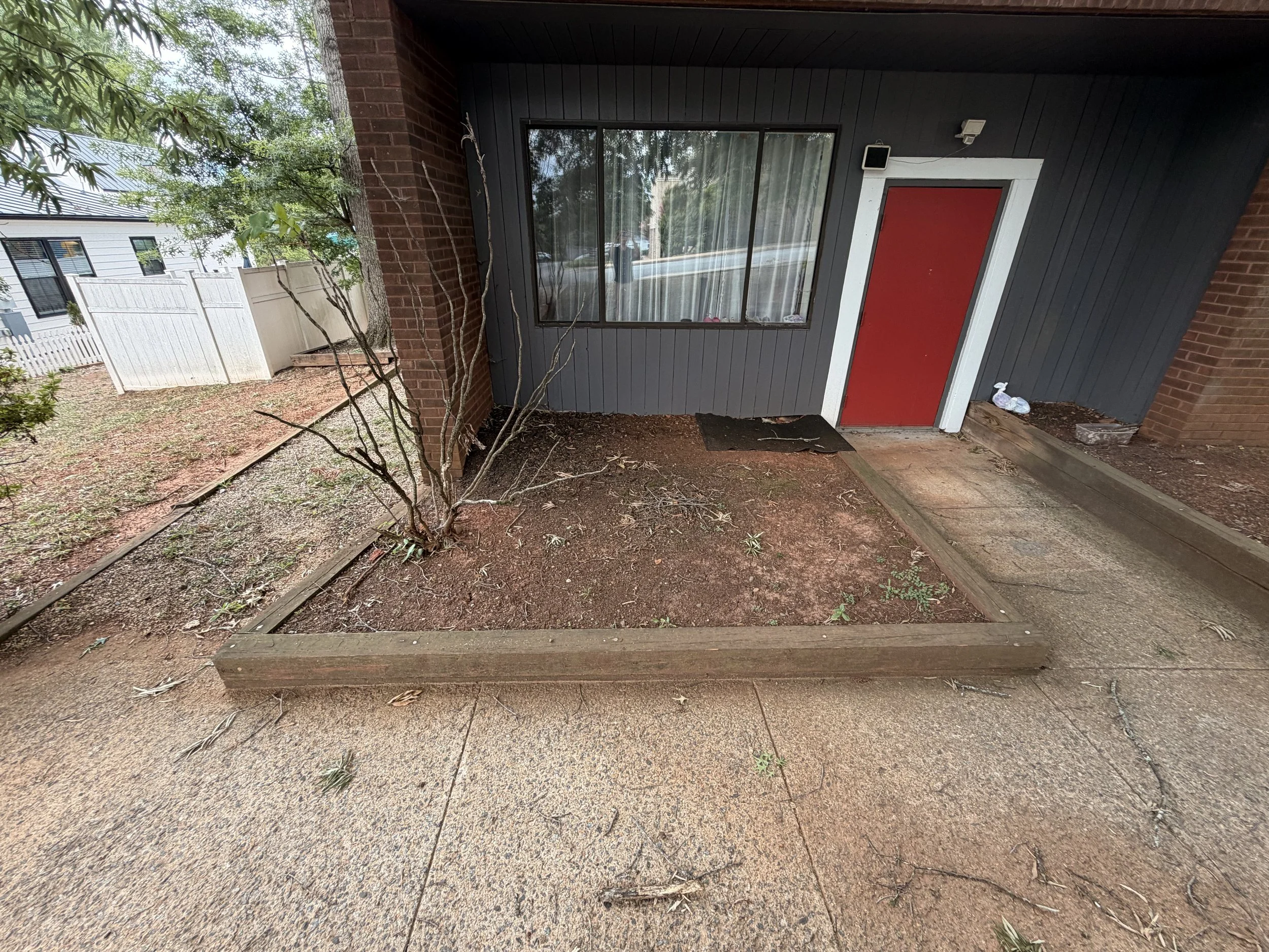View of a house entrance with a red door, gray exterior wall, and large window, surrounded by trees and a backyard with dirt and wooden borders.