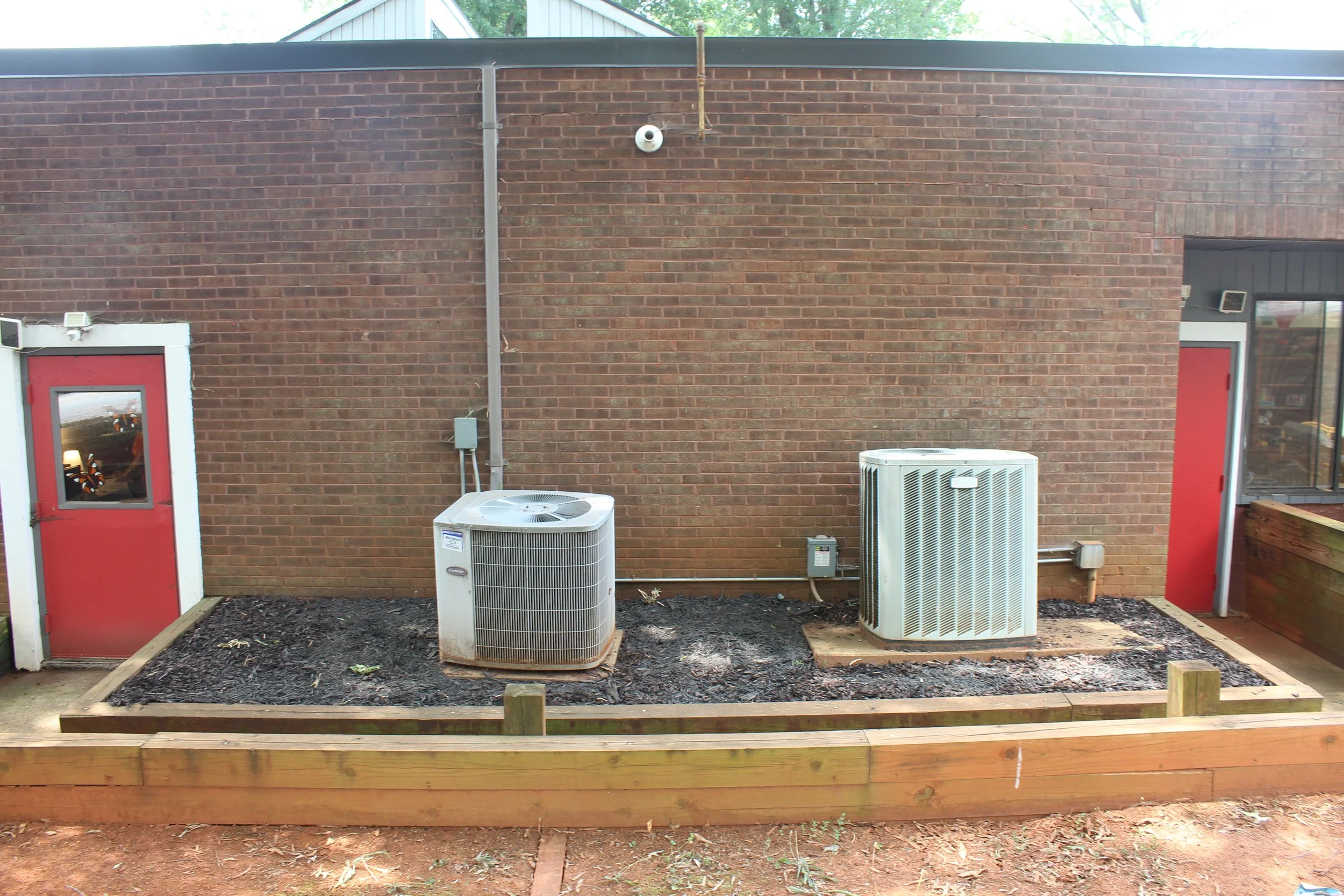 Backyard with two central air conditioning units on a black dirt patch, surrounded by a wooden frame. The background features a brick wall with two red doors, a small window, and visible pipes and electrical box.