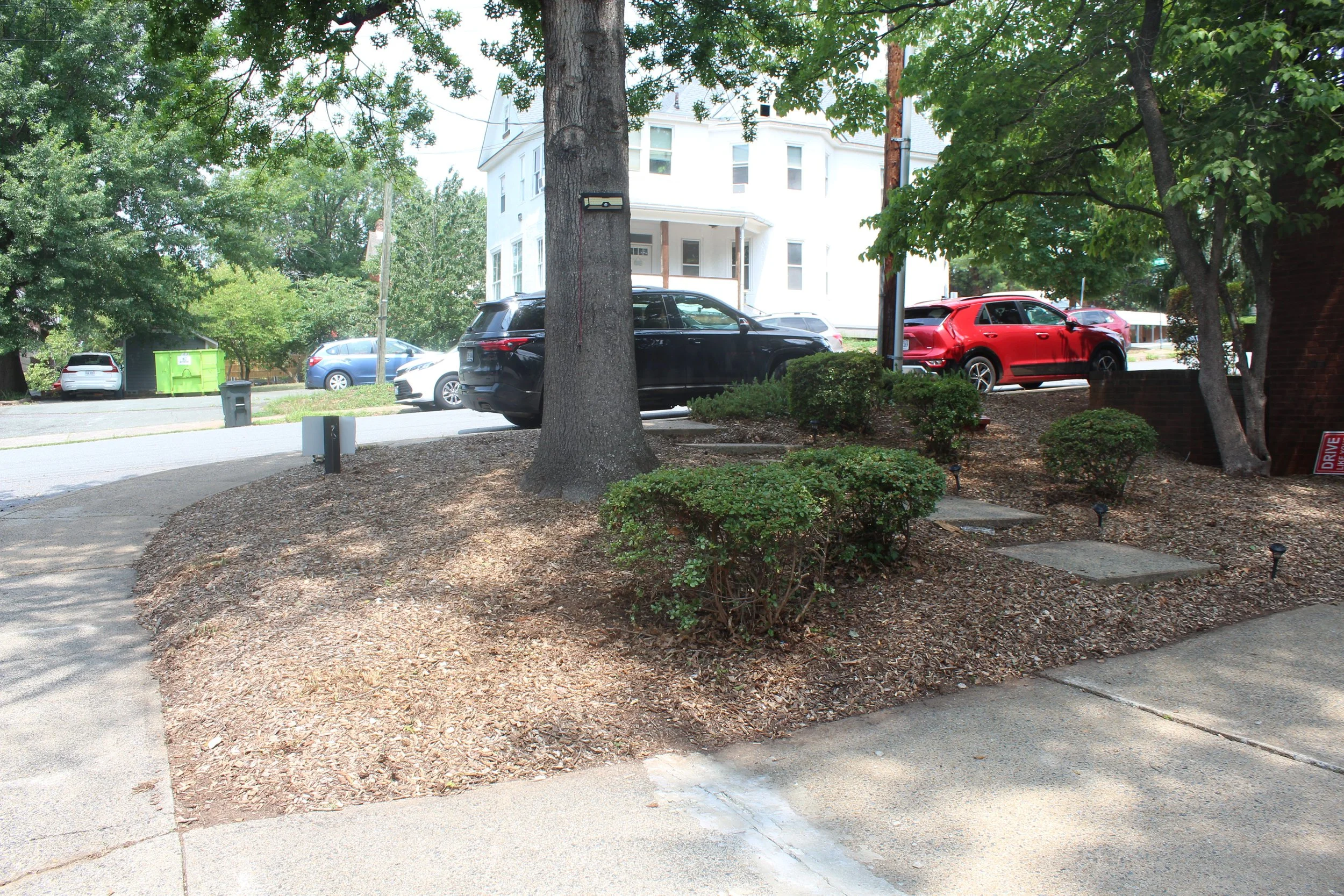 A residential street scene with trees, parked cars, and a white multi-story house in the background. The foreground features a landscaped area with small bushes and a sidewalk.