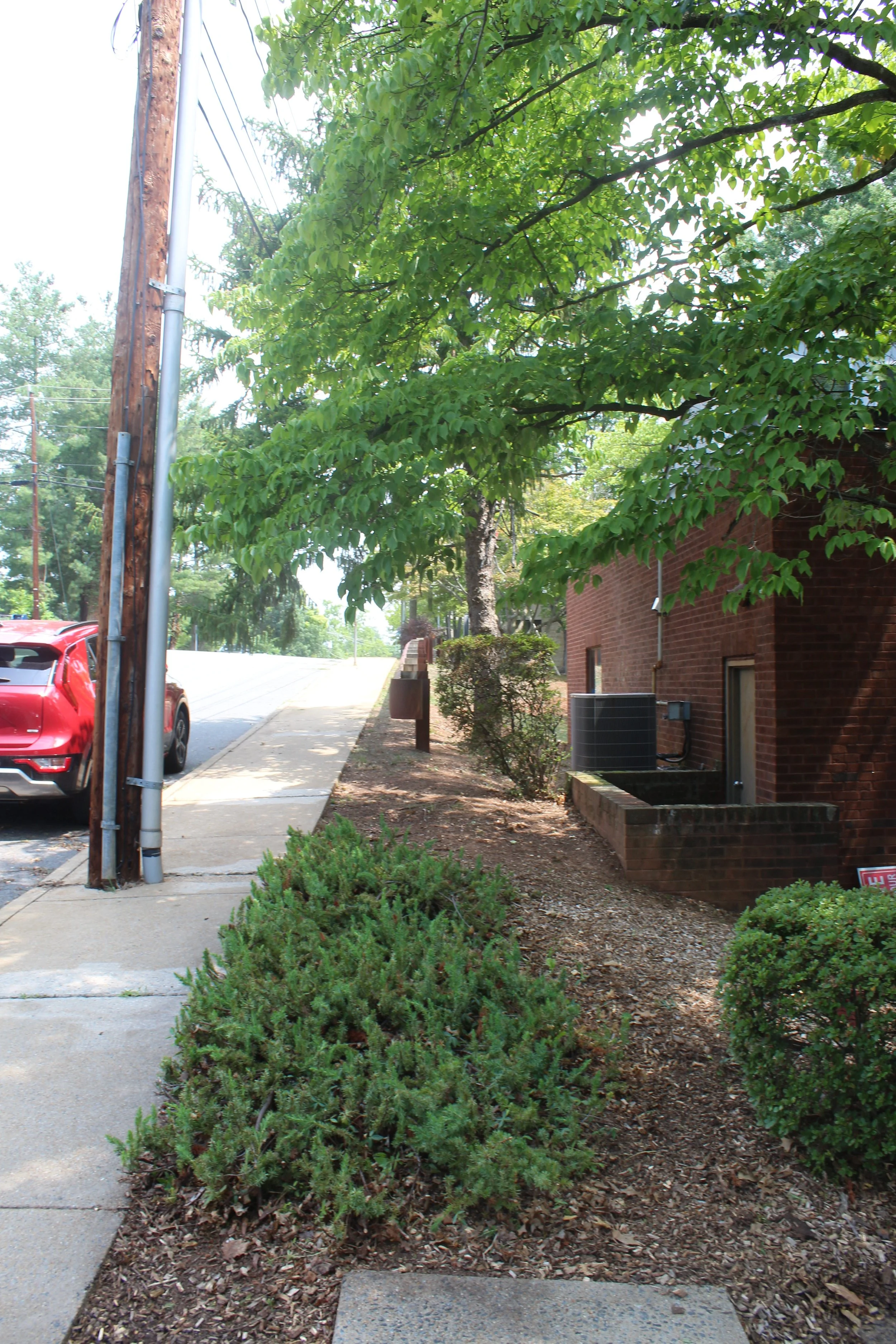 A sidewalk with bushes on the left side and a brick building on the right side. There are trees with green leaves overhead and a parked red car in the background. Utility poles and wires are visible, with some bushes and mulch along the sidewalk.