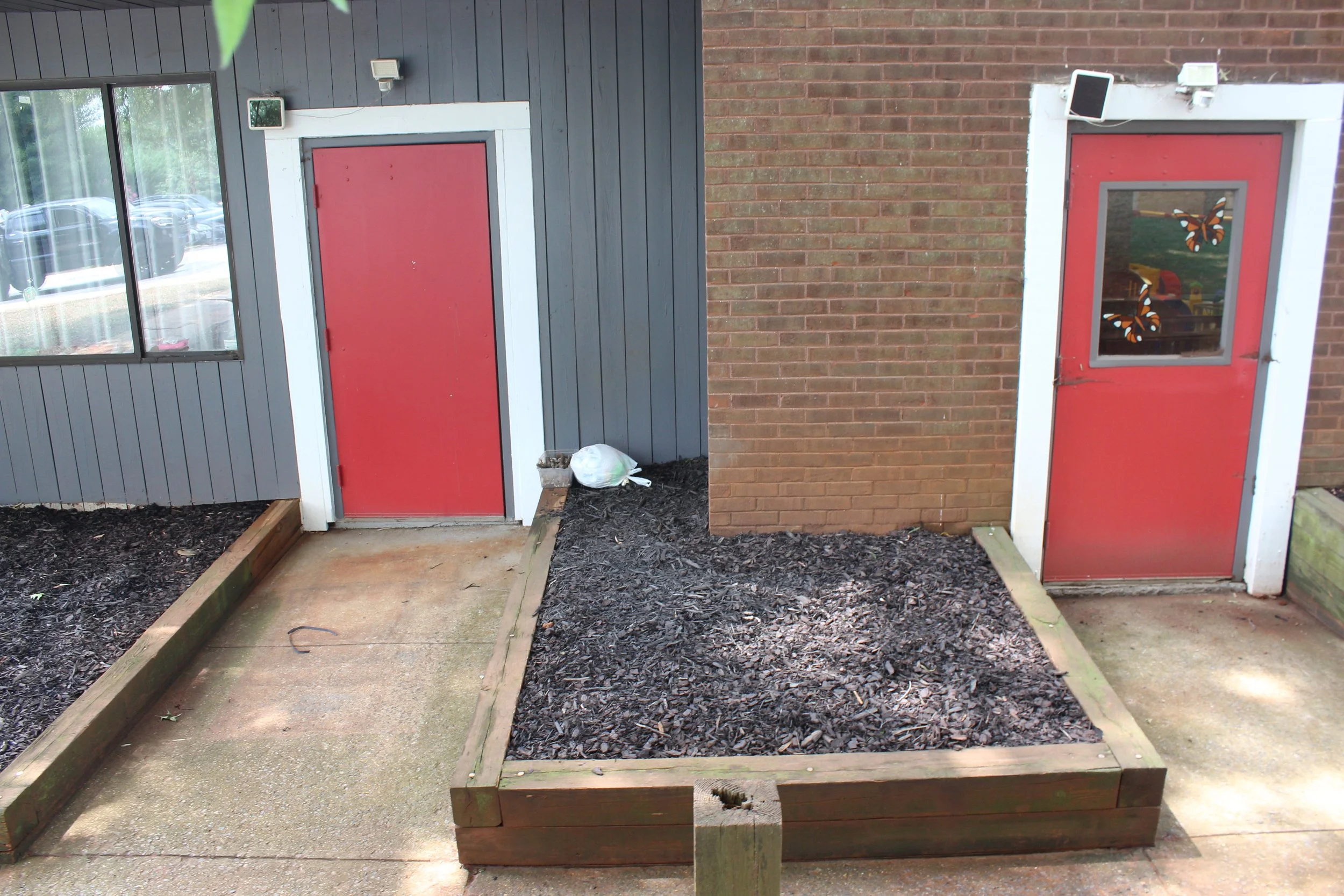 A garden bed with a wooden border filled with dark mulch, located in front of a building with a red door. To the left, there are large windows, and to the right, brick wall. A plastic bag and small planter are on the ground near the red door.