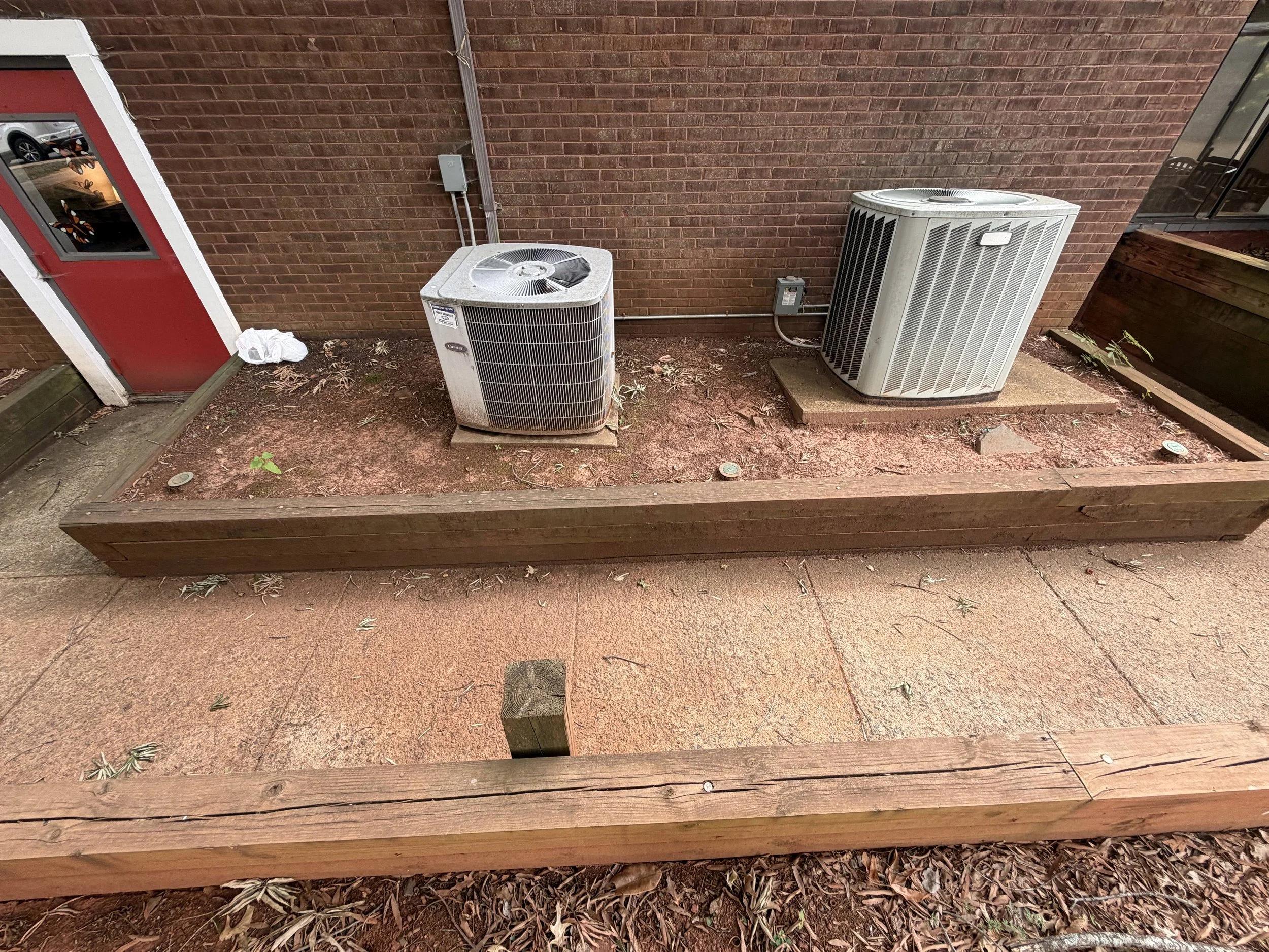 Two outdoor air conditioning units against a brick wall, with leaf debris and a wooden planter bed in the foreground.