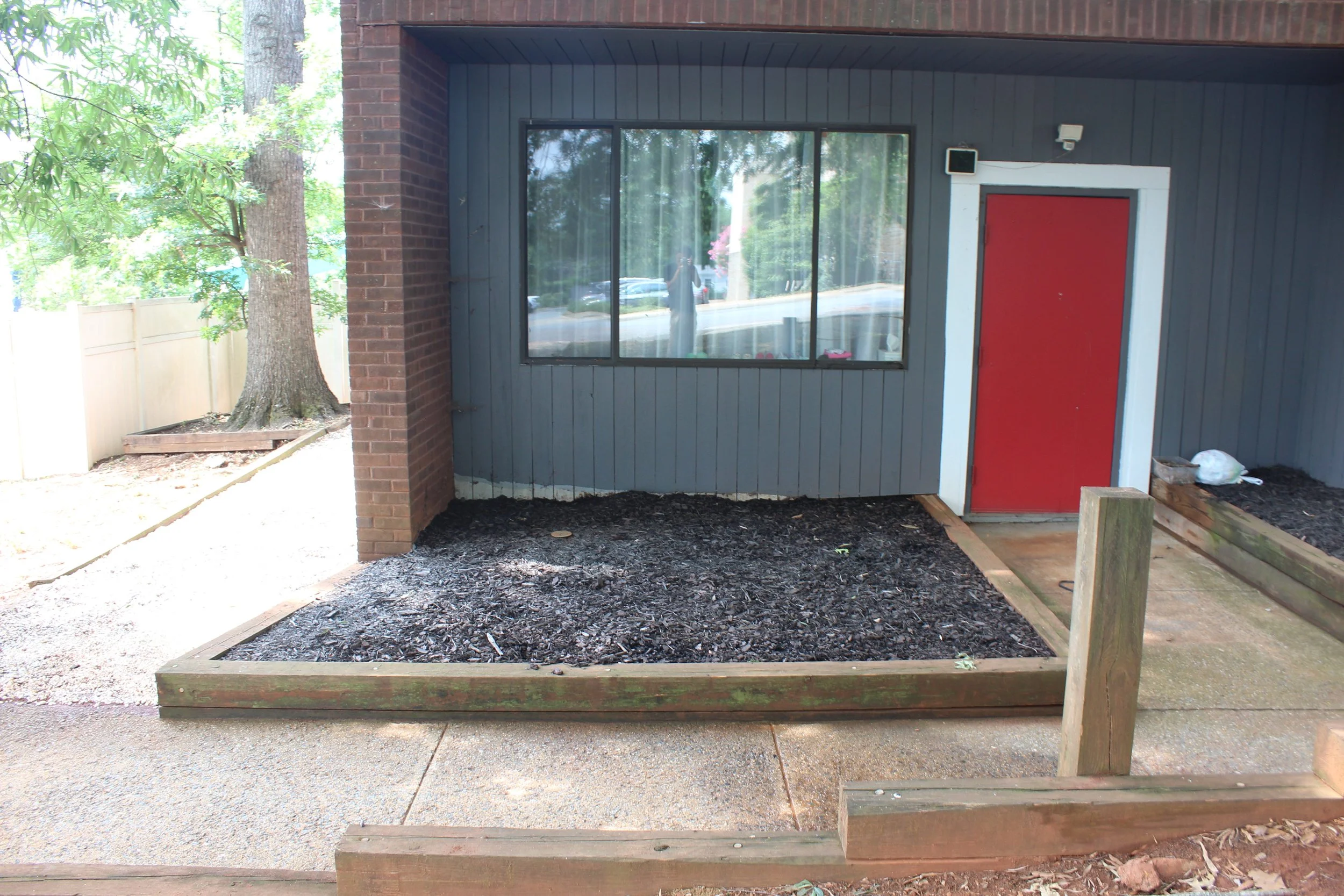 Backyard patio with a large window and red door. There is a tree and a wooden fence on the left, and a raised garden bed on the right with some gardening supplies. The ground is covered with mulch and concrete.