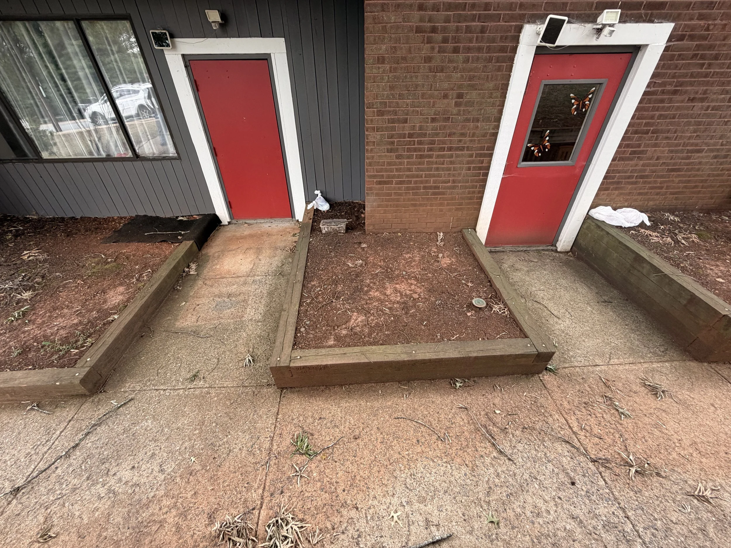 Red door with window, attached to a brick building, surrounded by wooden planter boxes, with a dirt and concrete pathway in front.